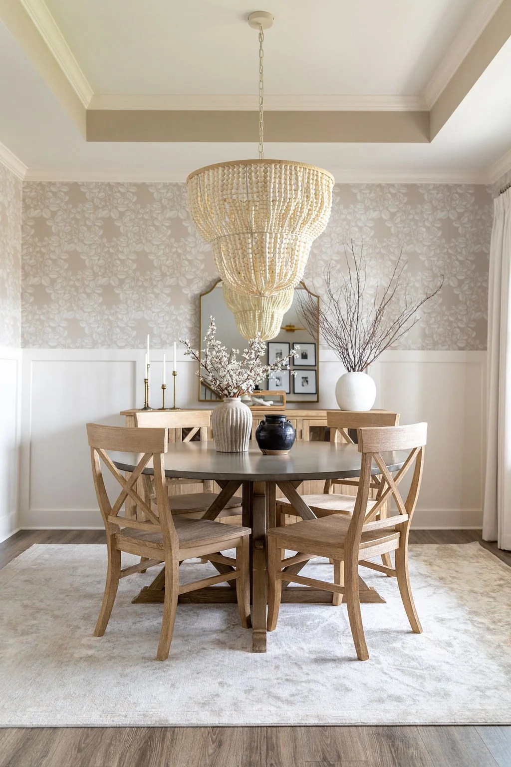 A dining room with a round wooden table, four matching wooden chairs, and decorative vases with branches and flowers. A large, multi-tiered chandelier hangs above the table. The room has light-colored walls with floral wallpaper, a mirror, framed pho
