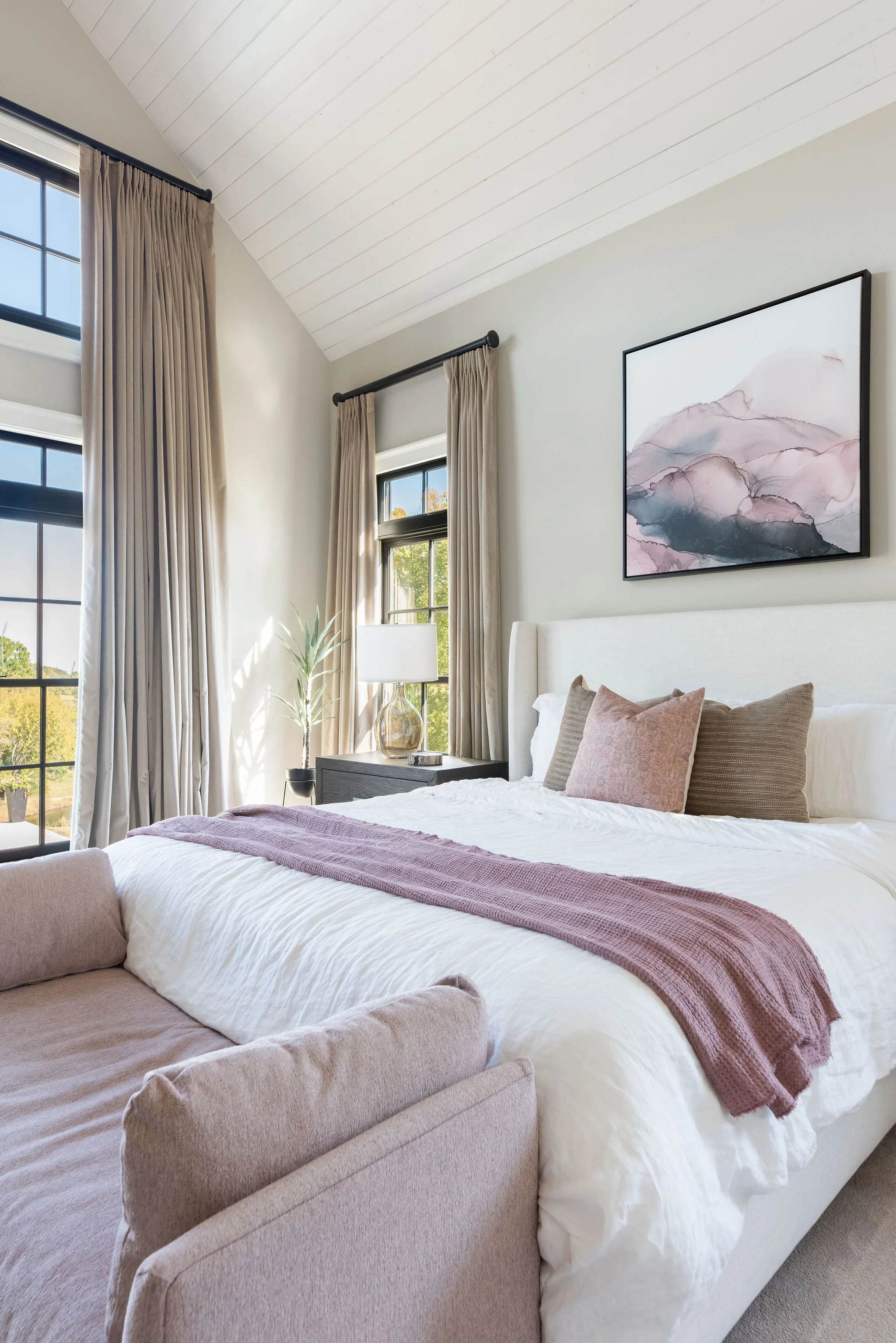 A cozy bedroom with a white bed, beige pillows, a pink throw blanket, a beige armchair, a nightstand with a glass lamp, and large windows with beige curtains.