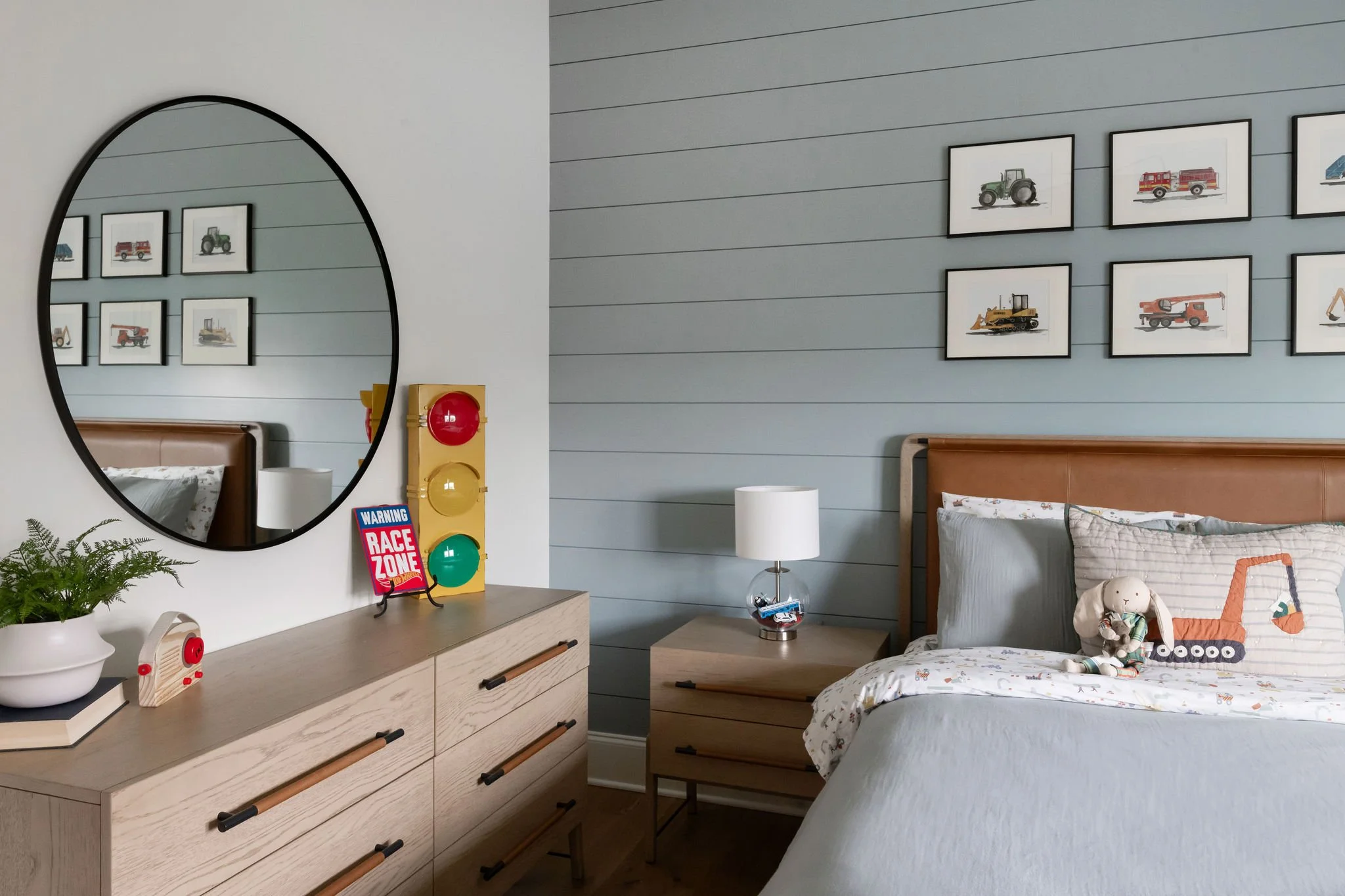 A bedroom with a blue shiplap accent wall, framed pictures of construction vehicles, a wooden dresser with a plant and kids' decor, a mirror, a nightstand with a lamp, and a bed with toys and bedding featuring construction equipment.