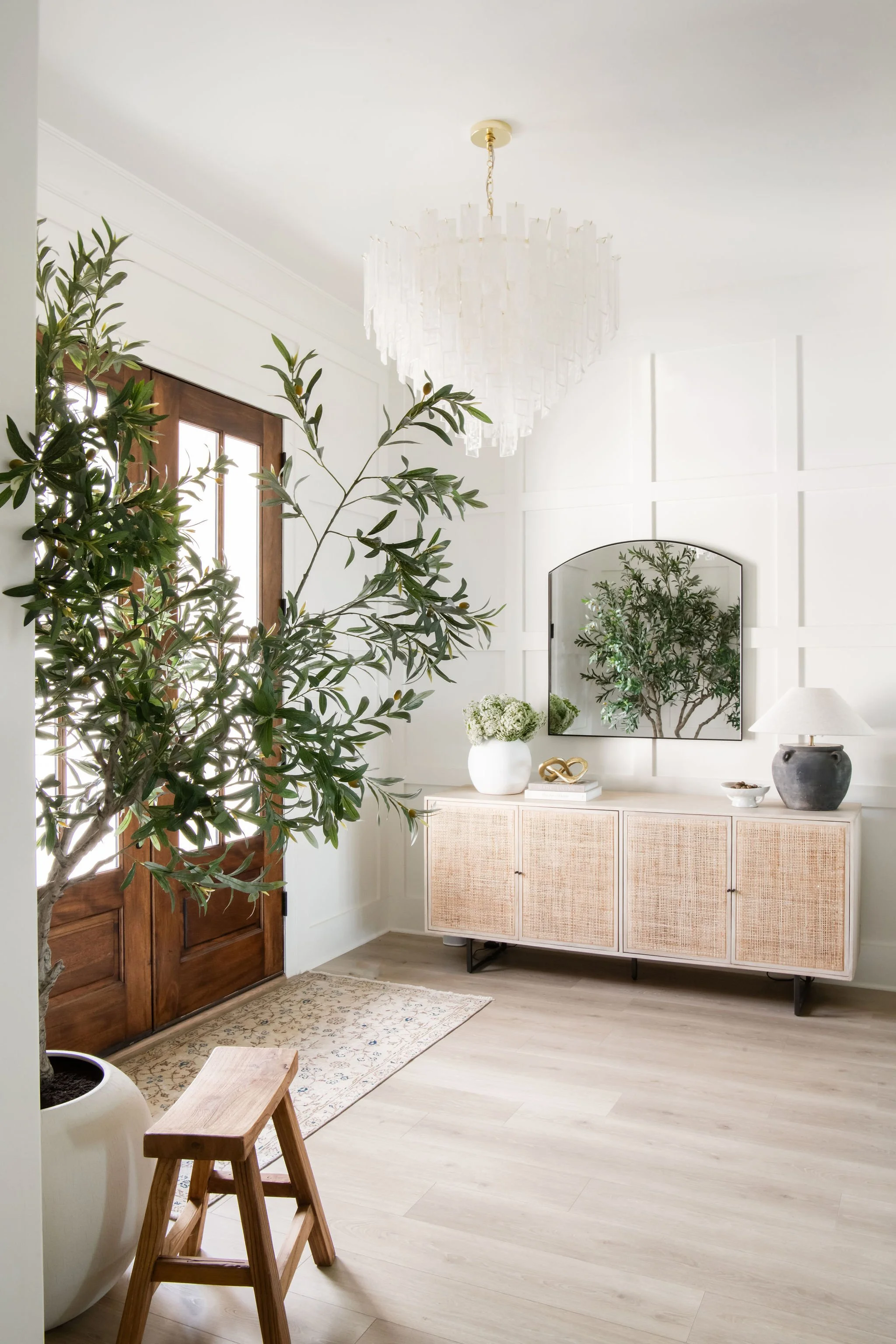 Bright entryway with large potted plant, wooden front door, white wall with paneling, modern sideboard with decorative items, mirror, lamp, and chandelier.