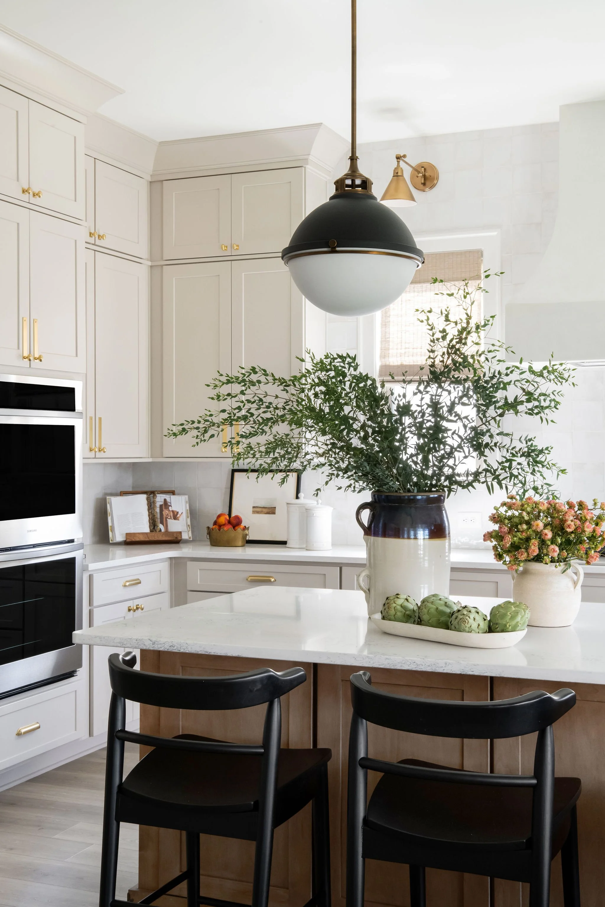 Modern kitchen with white cabinets, black and gold lighting fixtures, black chairs, and a large island with a white marble countertop, decorated with green artichokes and potted flowers.