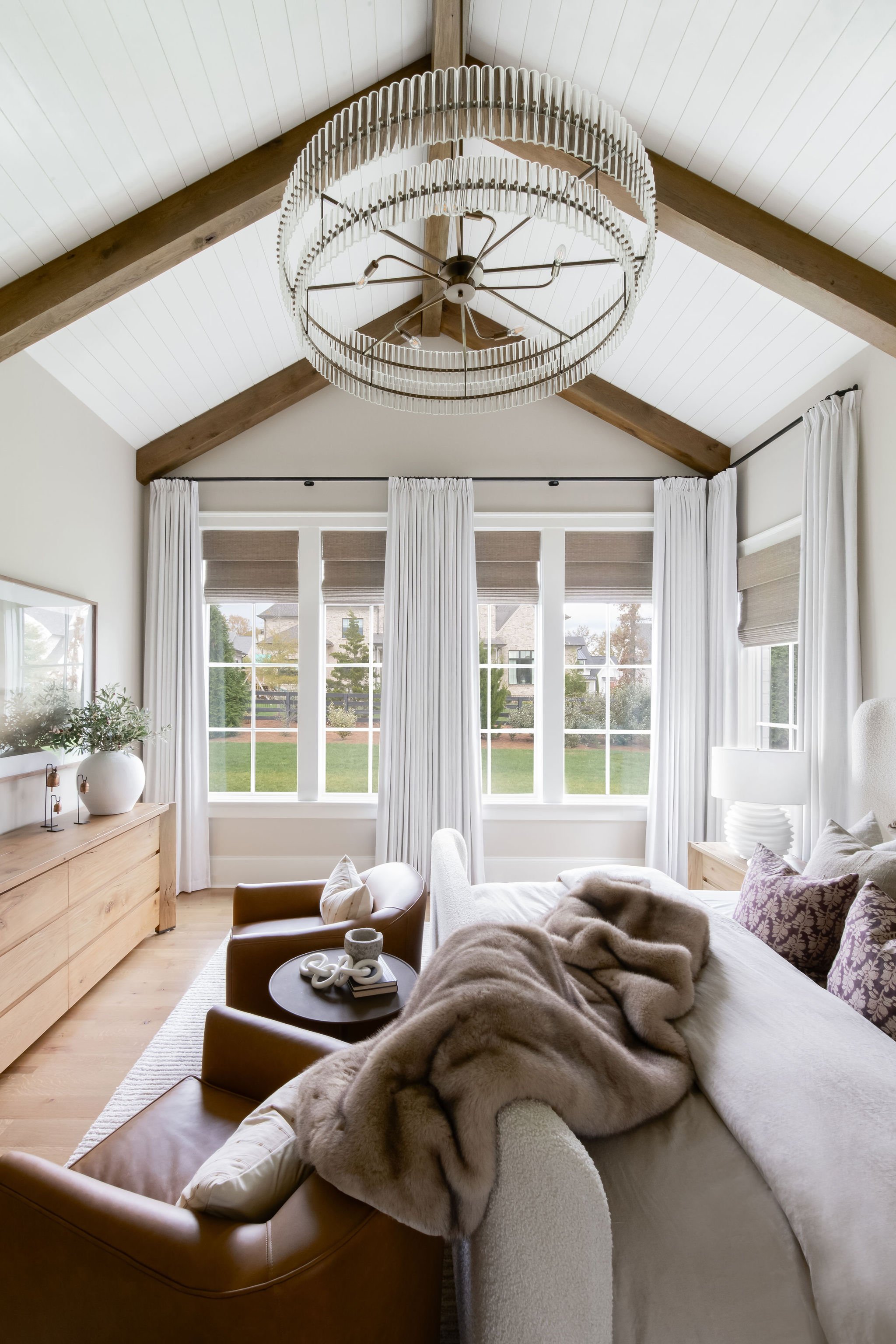 Living room with large window, white curtains, and vaulted ceiling with wooden beams, featuring a beige sofa with throw blanket, leather armchair, wooden dresser, and modern chandelier.
