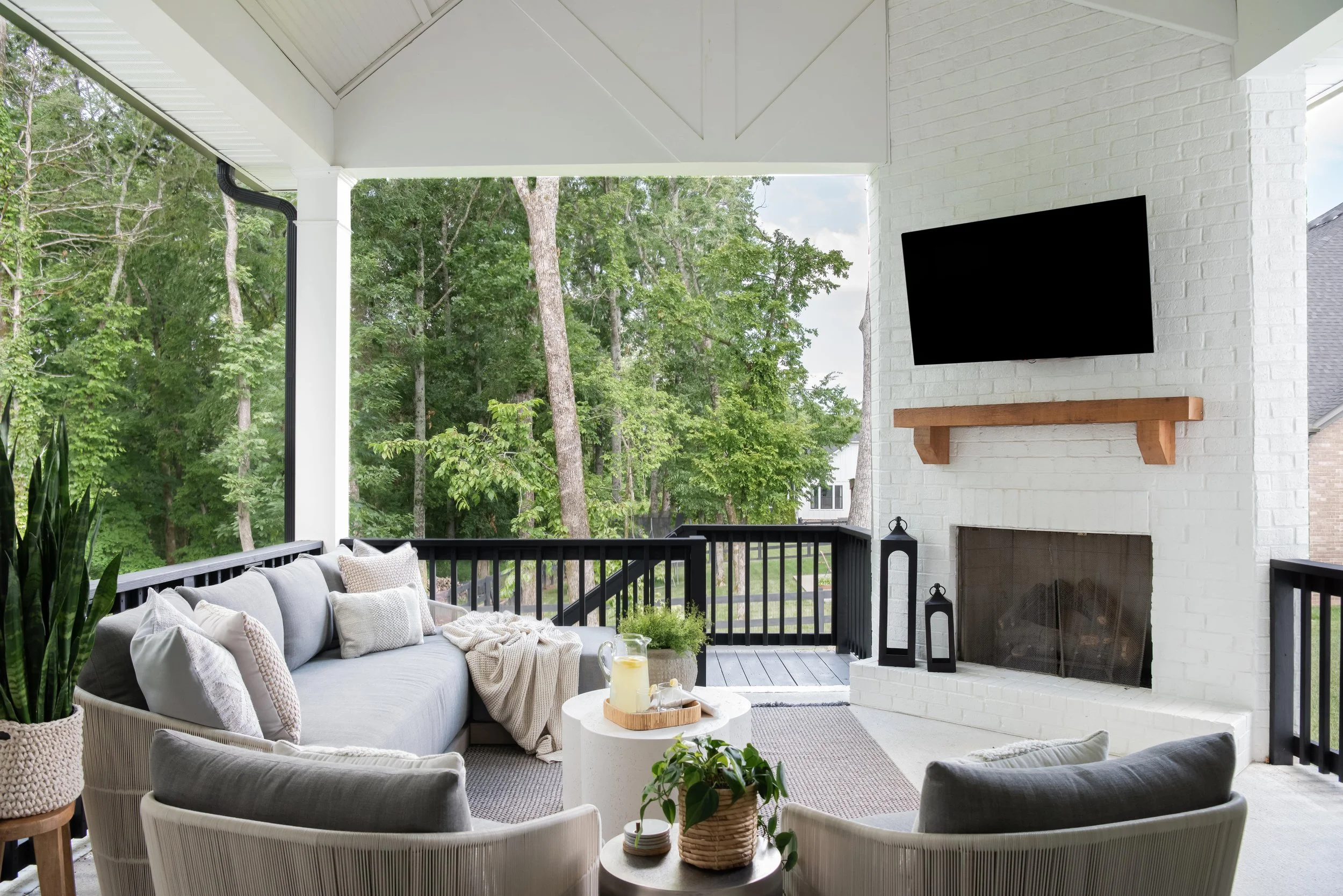 Outdoor covered porch with white brick fireplace, mounted flat-screen TV above a wooden mantel, and seating area with a gray sofa, white chairs, and small tables, surrounded by greenery and trees.