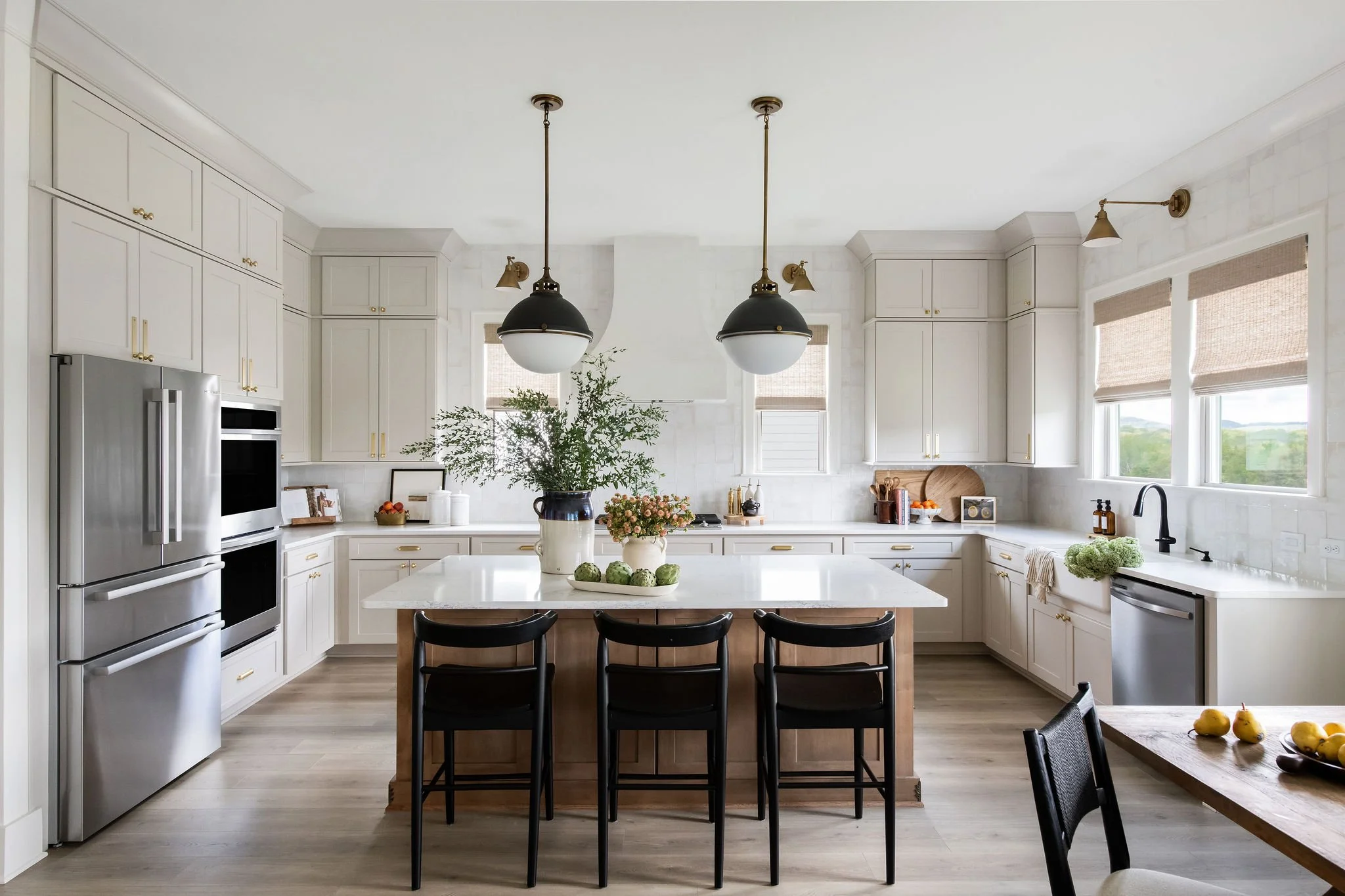 Bright, modern kitchen with white cabinets, stainless steel appliances, a central island with black chairs, and large windows with shades.
