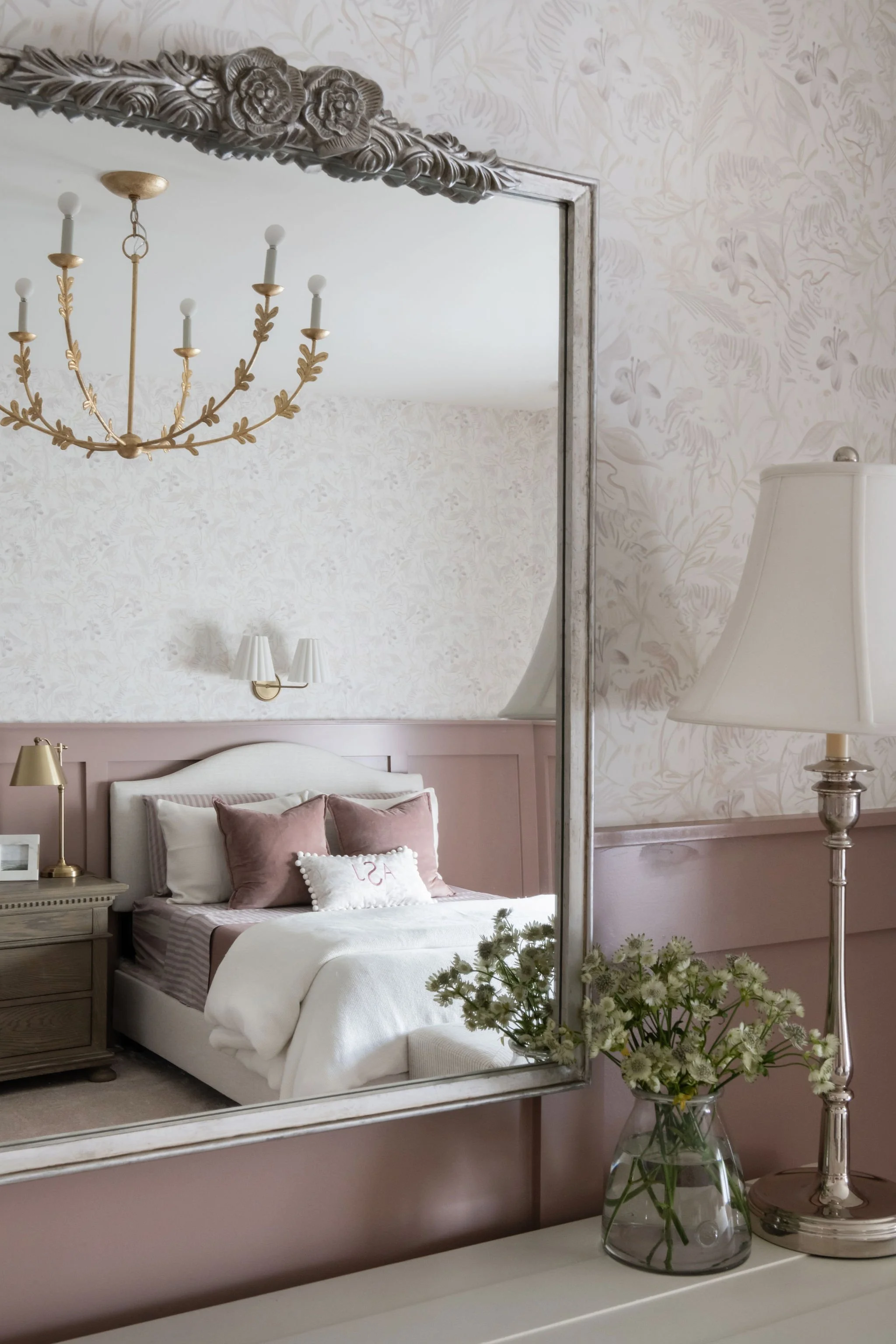 A bedroom reflected in a mirror with a chandelier and wall-mounted lights, pink and white bedding, a nightstand with a lamp, and a vase of white flowers.