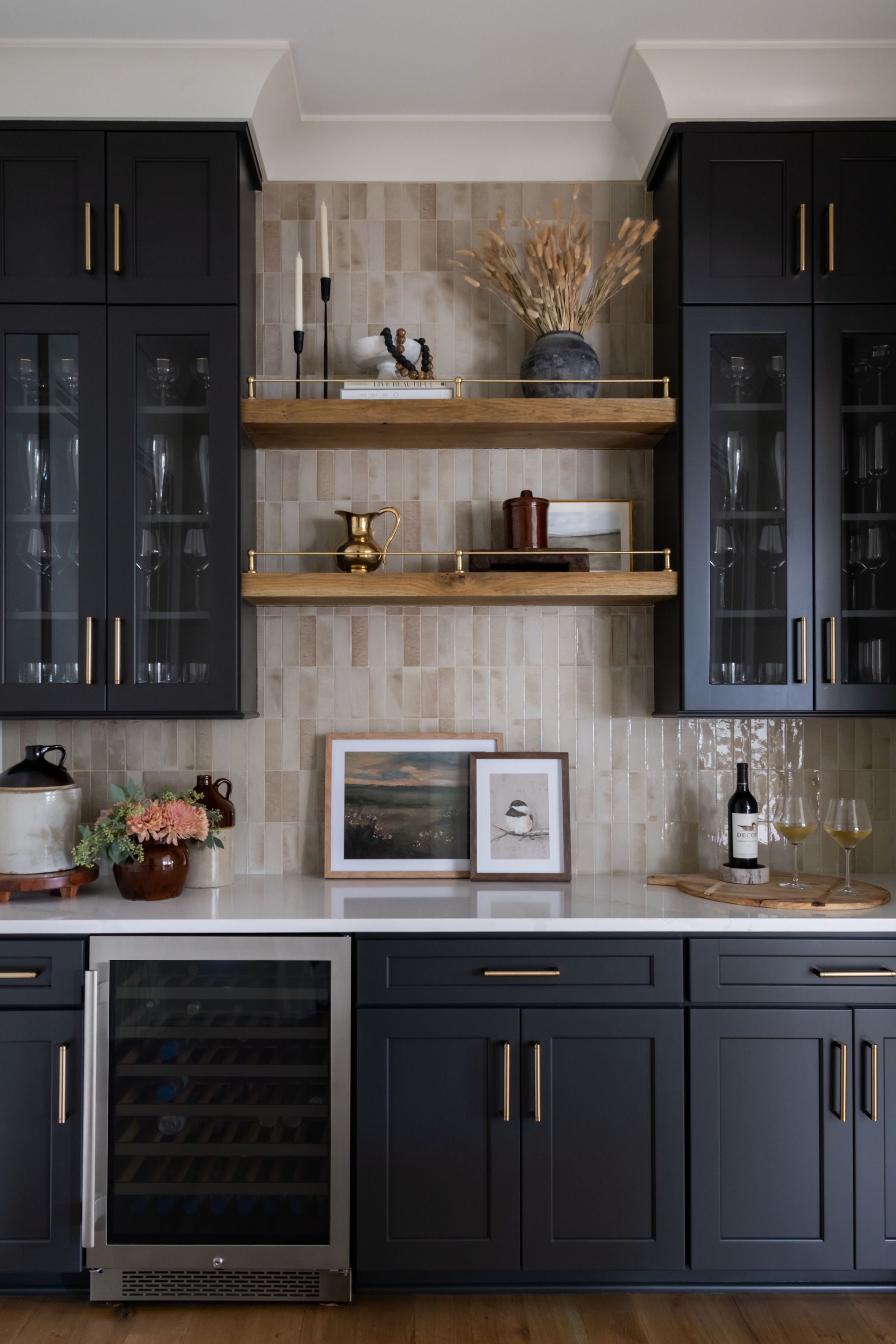 Modern kitchen with black cabinets, glass-front upper cabinets, and open wooden shelves decorated with vases, candles, artwork, and wine glasses.