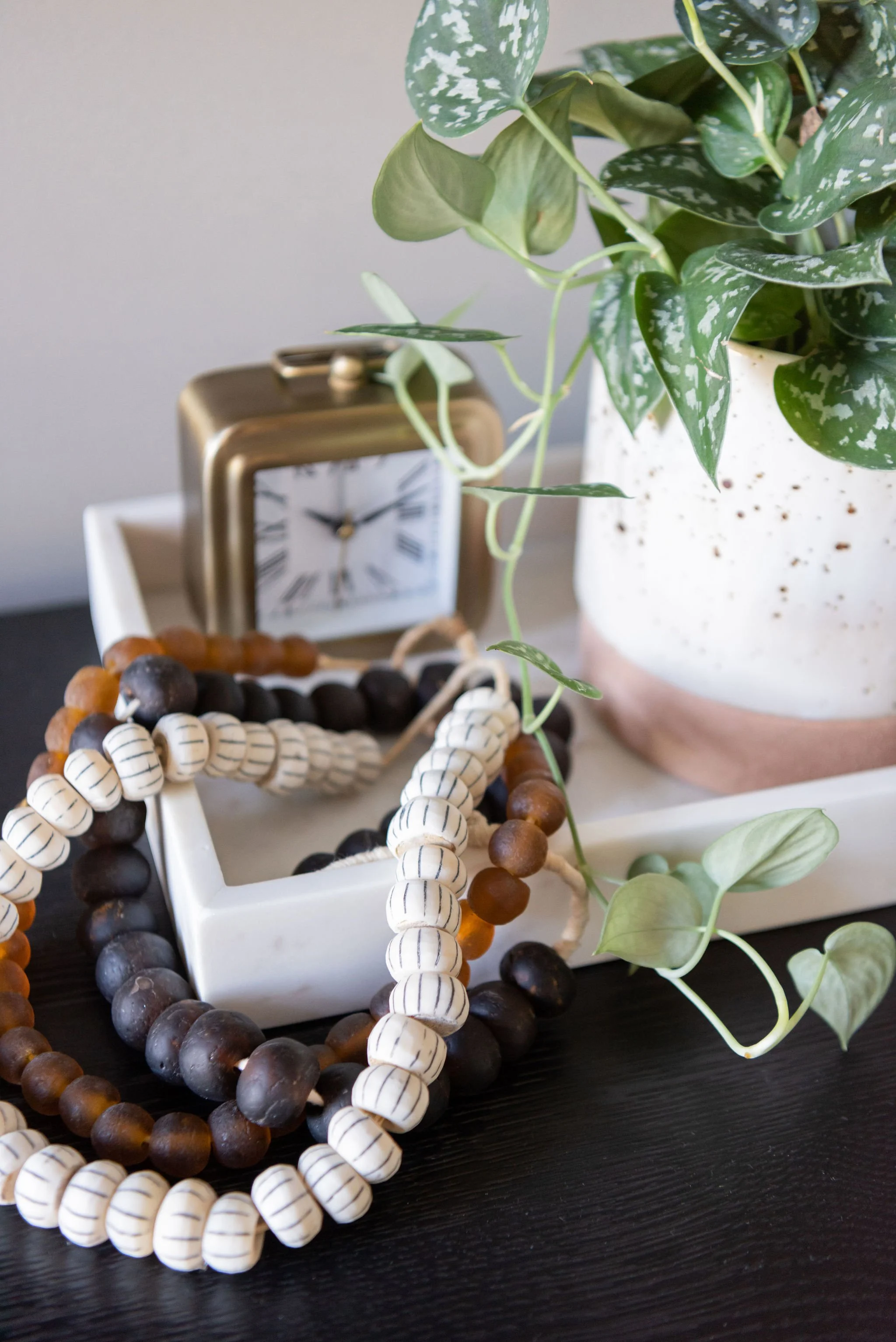 Decorative objects on a black surface including a gold alarm clock, a white tray with wooden beaded jewelry, a speckled white and pink planter with green leafy plant, and a small white tray.