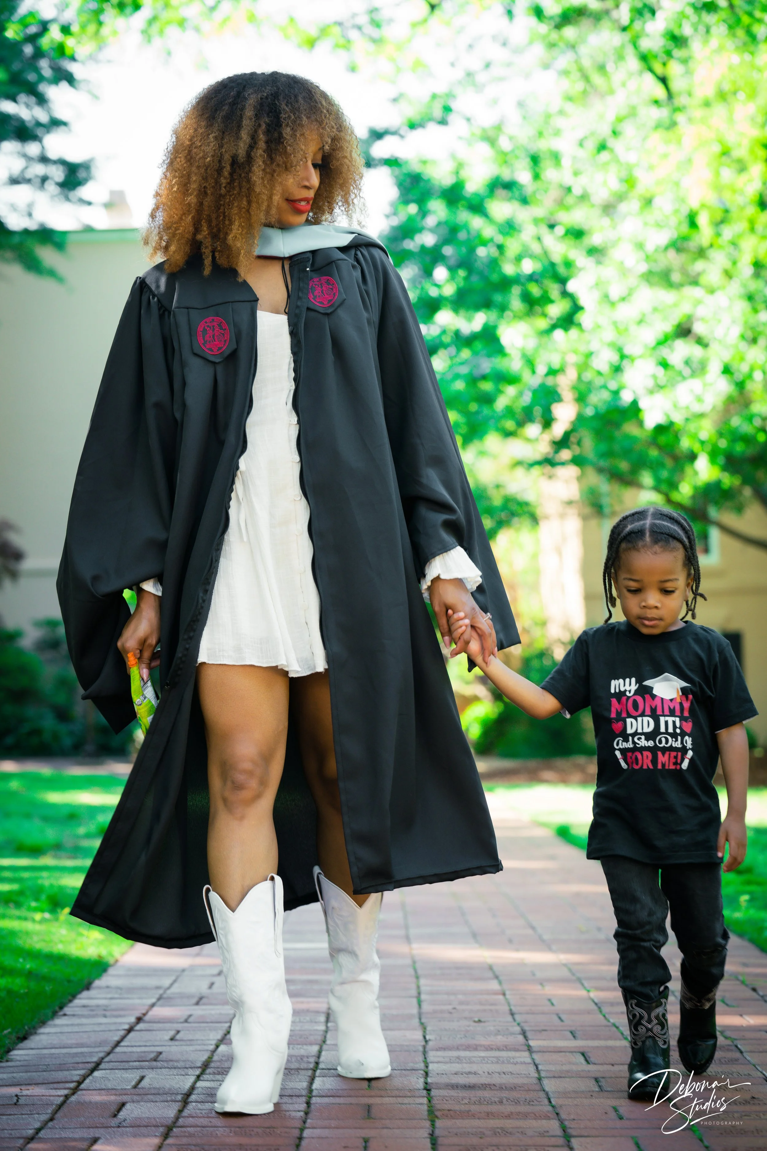 A woman in a graduation gown holding hands with a young girl wearing a black t-shirt with pink and white writing and black pants, walking on a brick sidewalk in a lush green outdoor setting.