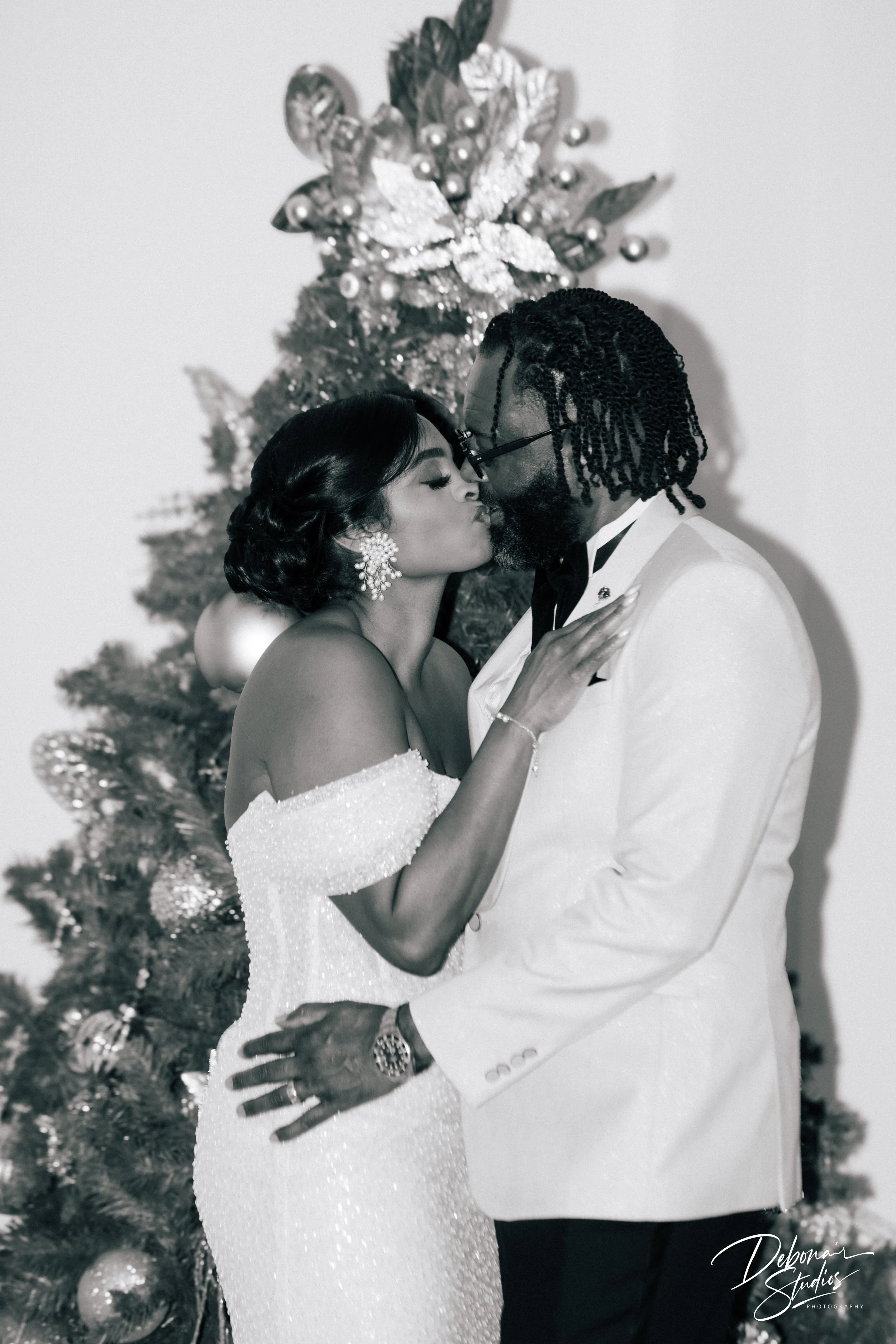 A black-and-white photo of a couple in formal attire sharing an intimate moment in front of a decorated Christmas tree, with the woman in an off-shoulder gown and the man in a white tuxedo.