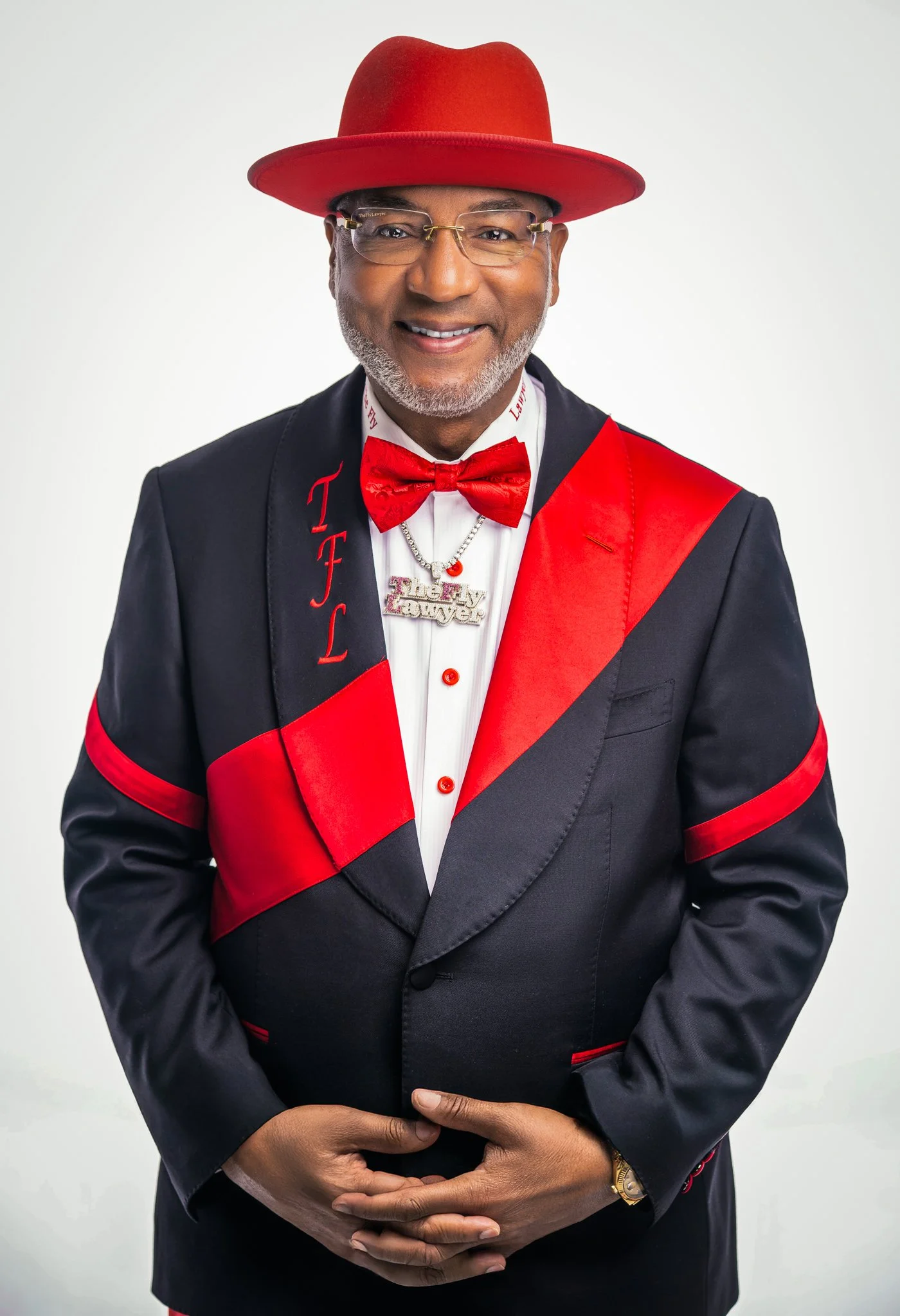 A man dressed in a black tuxedo with red accents, wearing glasses, a red hat, red bow tie, and a necklace that says 'Thelk Lawler', standing against a white background.