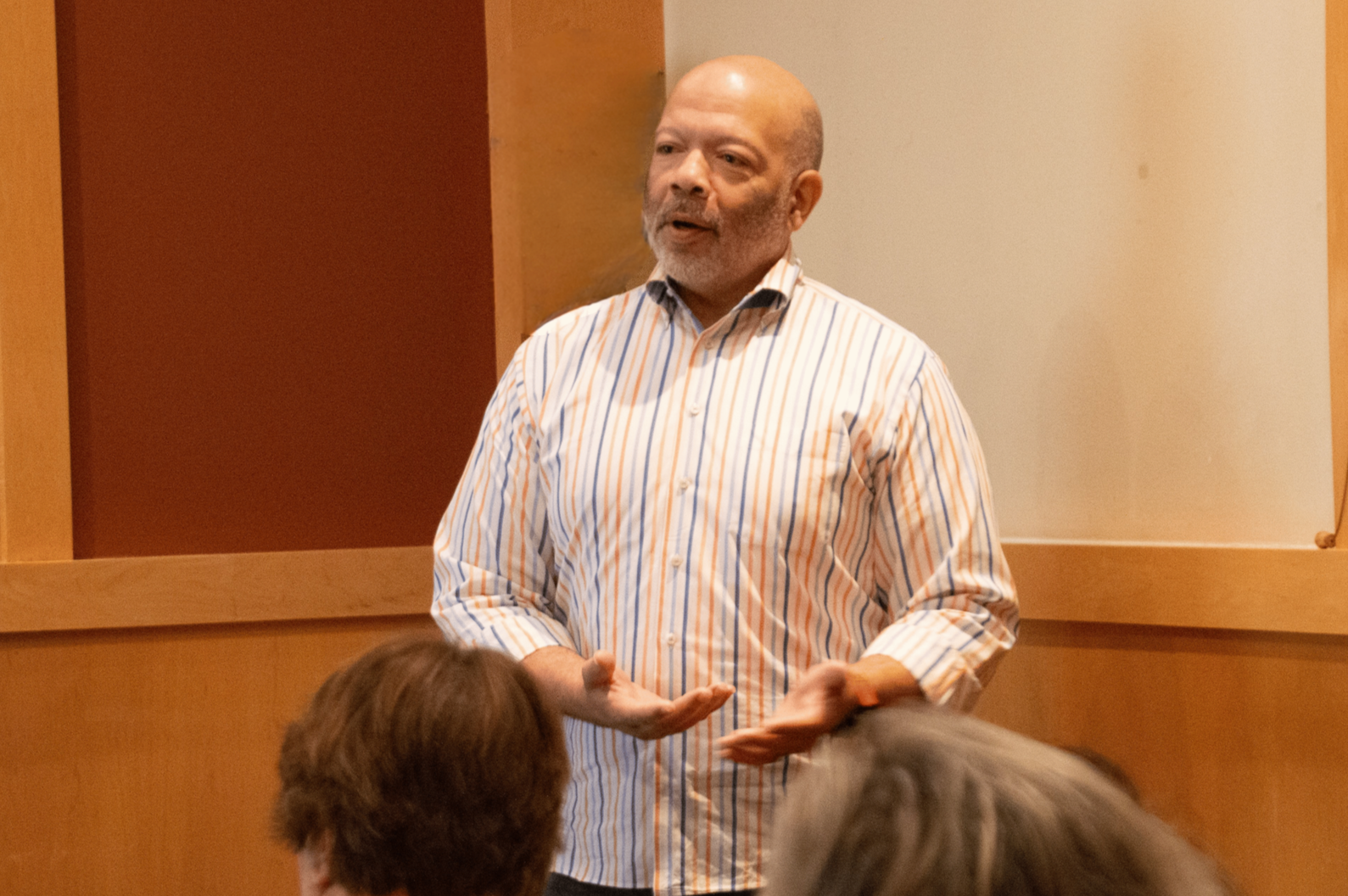 Man with a bald head and gray beard speaking to a group of people in a room with wood paneling and white walls, gesturing with his hands.