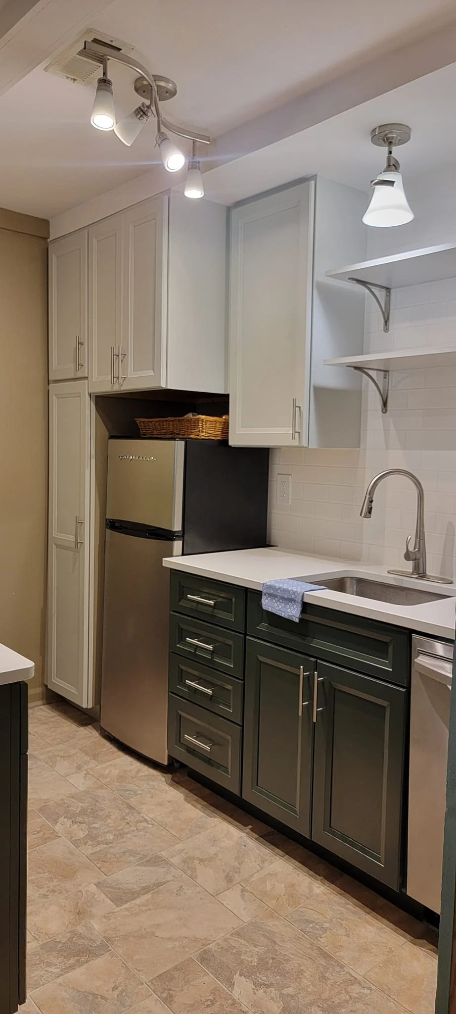 Kitchen with upper white cabinets, lower dark green cabinets, a small refrigerator, open shelves, a single basin sink with a modern faucet, a blue cloth on the countertop, and tiled floor.