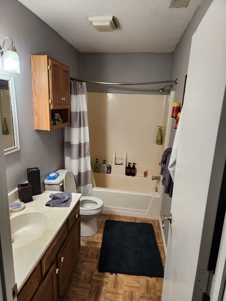 A small bathroom with a single sink vanity, wooden cabinets, a mirror, a window, a toilet, a bathtub with shower curtain, and various toiletries on the ledge and shelves.