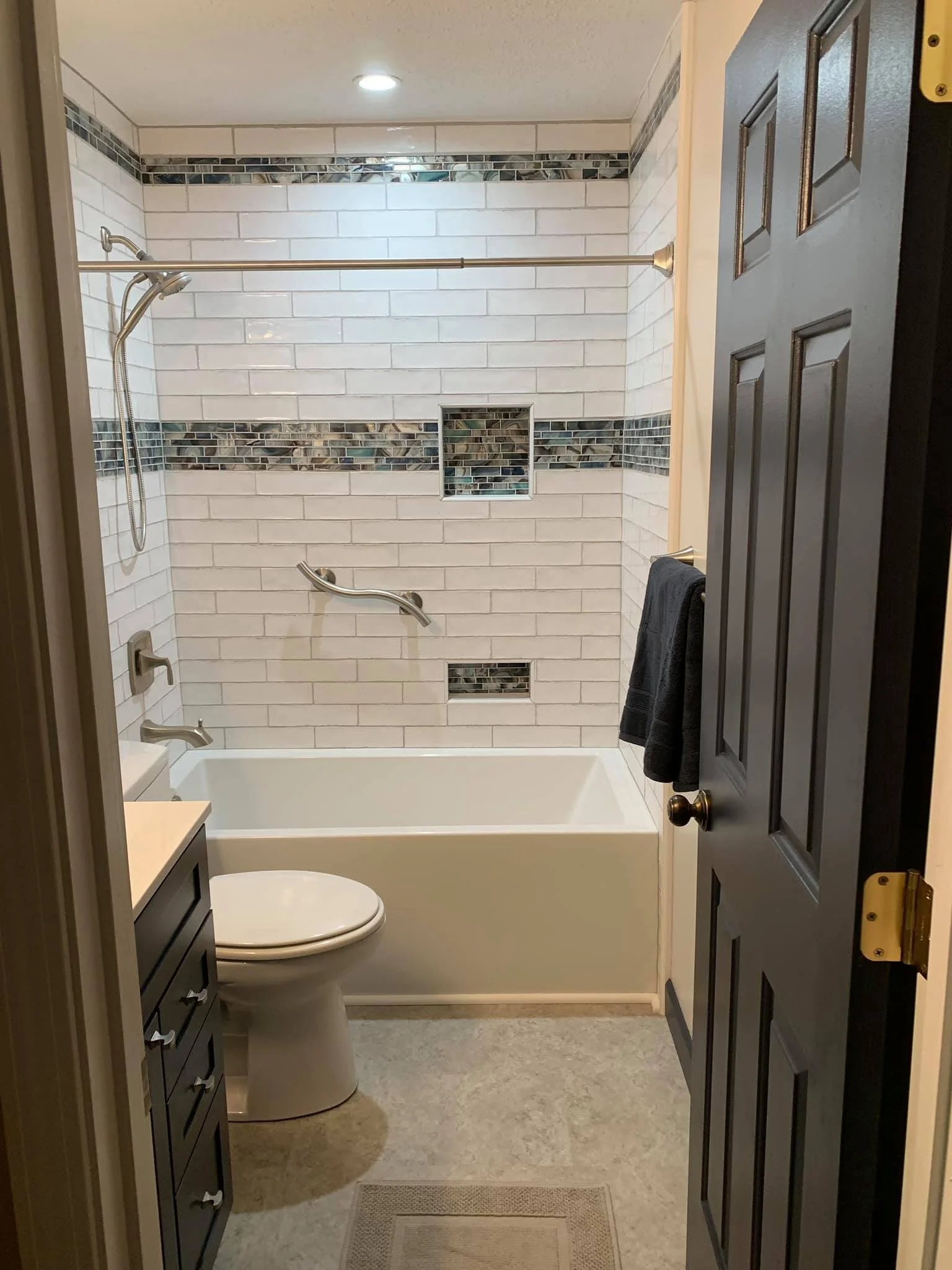 A bathroom shower with white subway tiles, a decorative tile border, built-in shelves, a showerhead, and a grab bar. There is a toilet and cabinetry visible in the foreground and a partially open dark wood door.