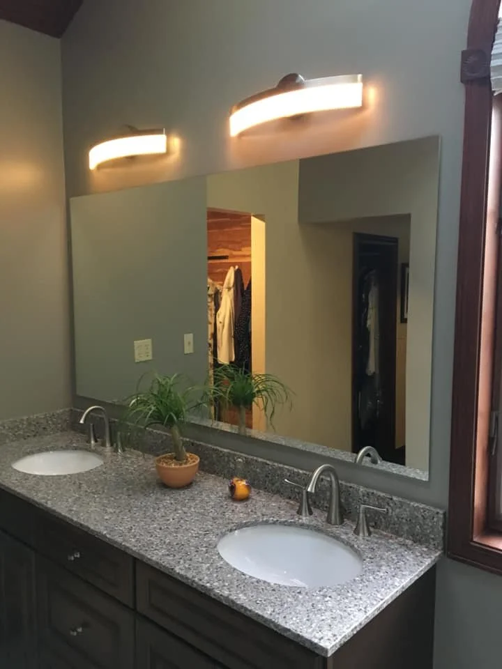 A bathroom vanity with a granite countertop, two sinks, and a large mirror. Two modern light fixtures are mounted above the mirror, and a potted plant with two green plants is on the countertop. There is also a small decorative frog figurine.