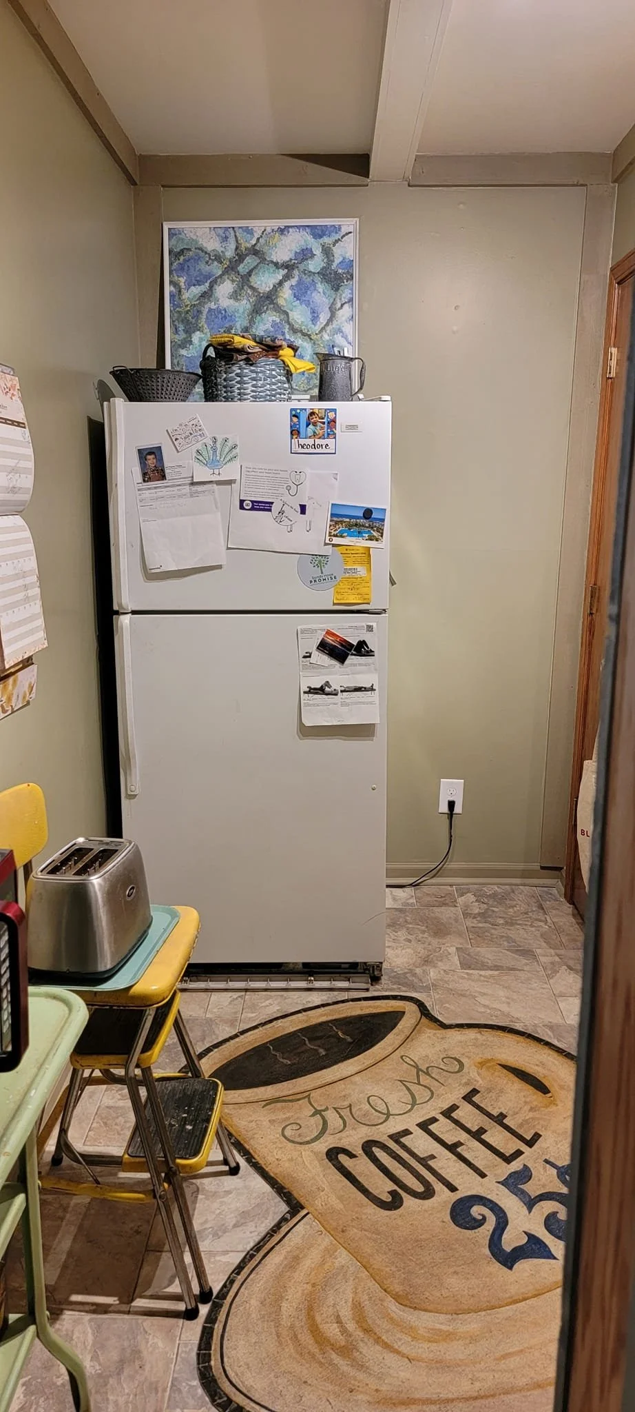 A white refrigerator with various magnets and papers on the front, located in a kitchen with beige walls, a floor mat that says 'Fresh Coffee,' a toaster, small chairs, and a painting on the wall above the fridge.