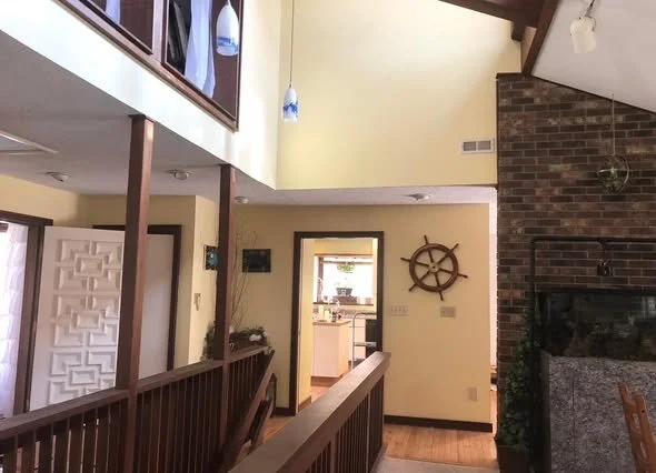 Interior of a house with a brick fireplace, a ship wheel decor, and a view into the kitchen.