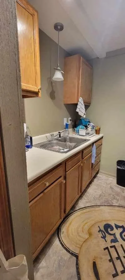 Kitchen sink area with wooden cabinets, a hanging light, and various kitchen items on the countertop, including dish soap and a towel.