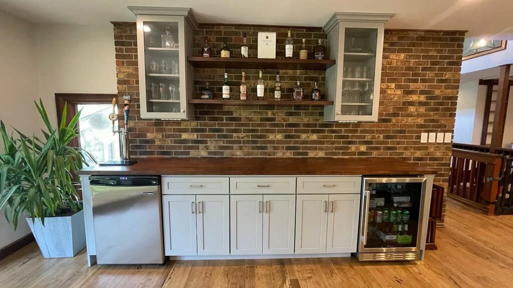 A home bar area with white cabinets, a wooden countertop, a mini fridge, a wine fridge, and a brick backsplash with shelves holding liquor bottles.