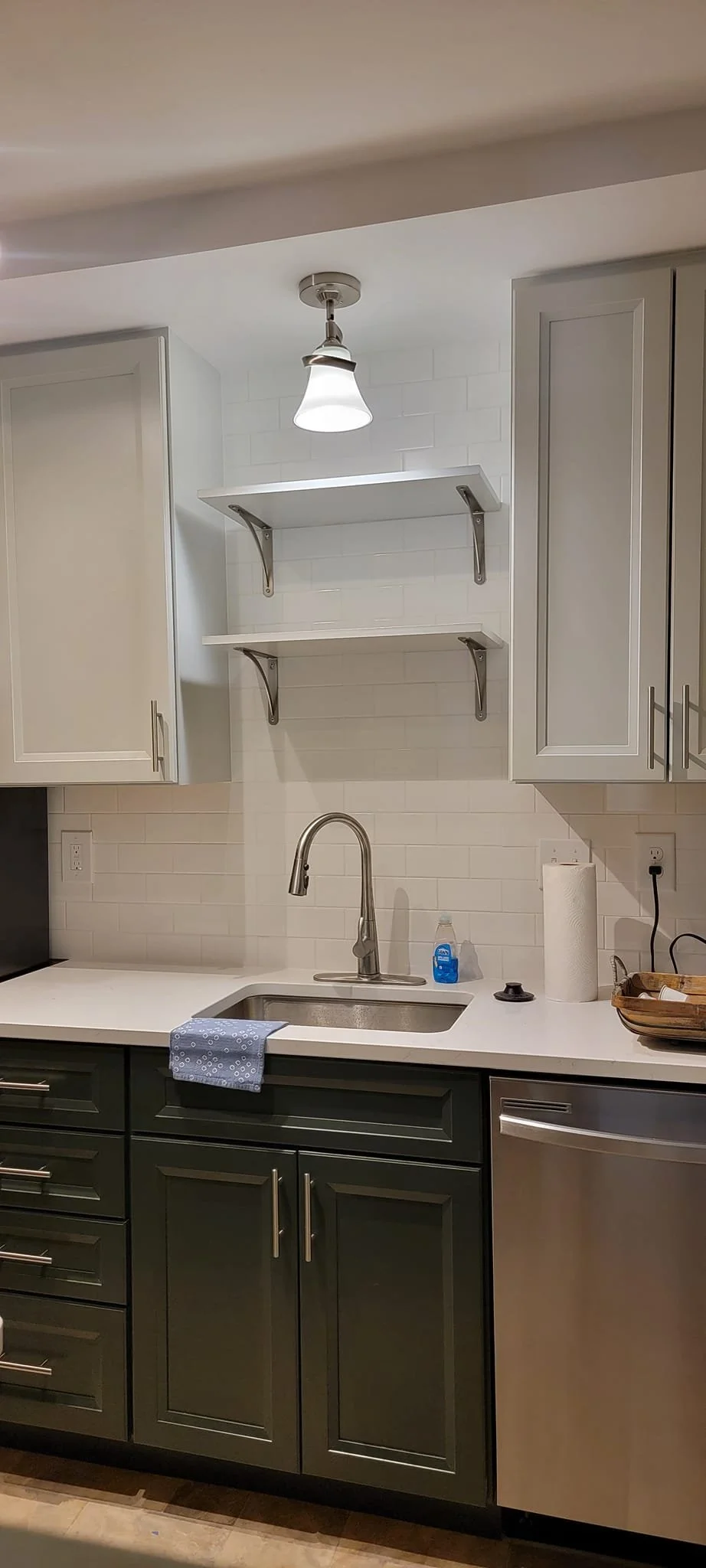 Kitchen with dark green lower cabinets, white countertop, a stainless steel sink with a curved faucet, two open white shelves tied with metallic brackets, white backsplash tiles, and a paper towel roll. There is a small cleaning spray, a black lid, a