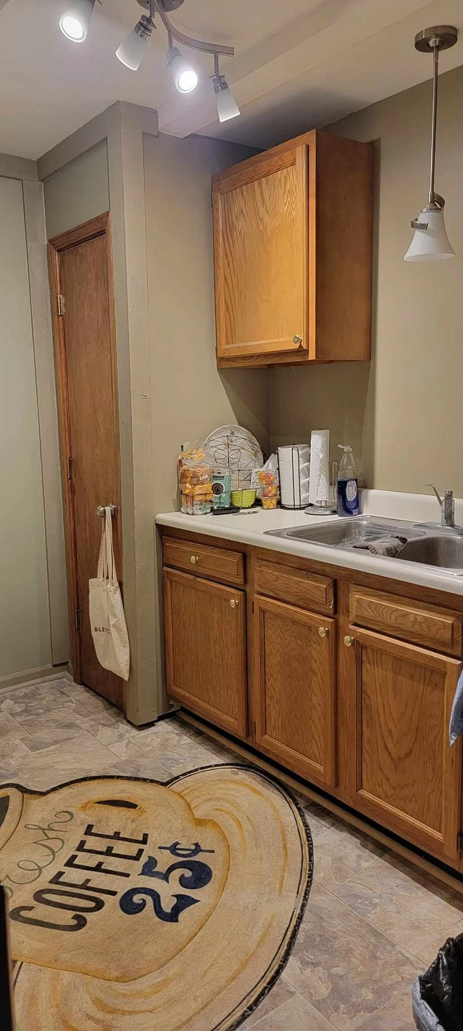 Kitchen with wooden cabinets, a white countertop, and a sink. Items on the counter include a dish rack, paper towels, and soap. On the floor, there is a coffee-themed rug with the text 'Coffee $2.50'.