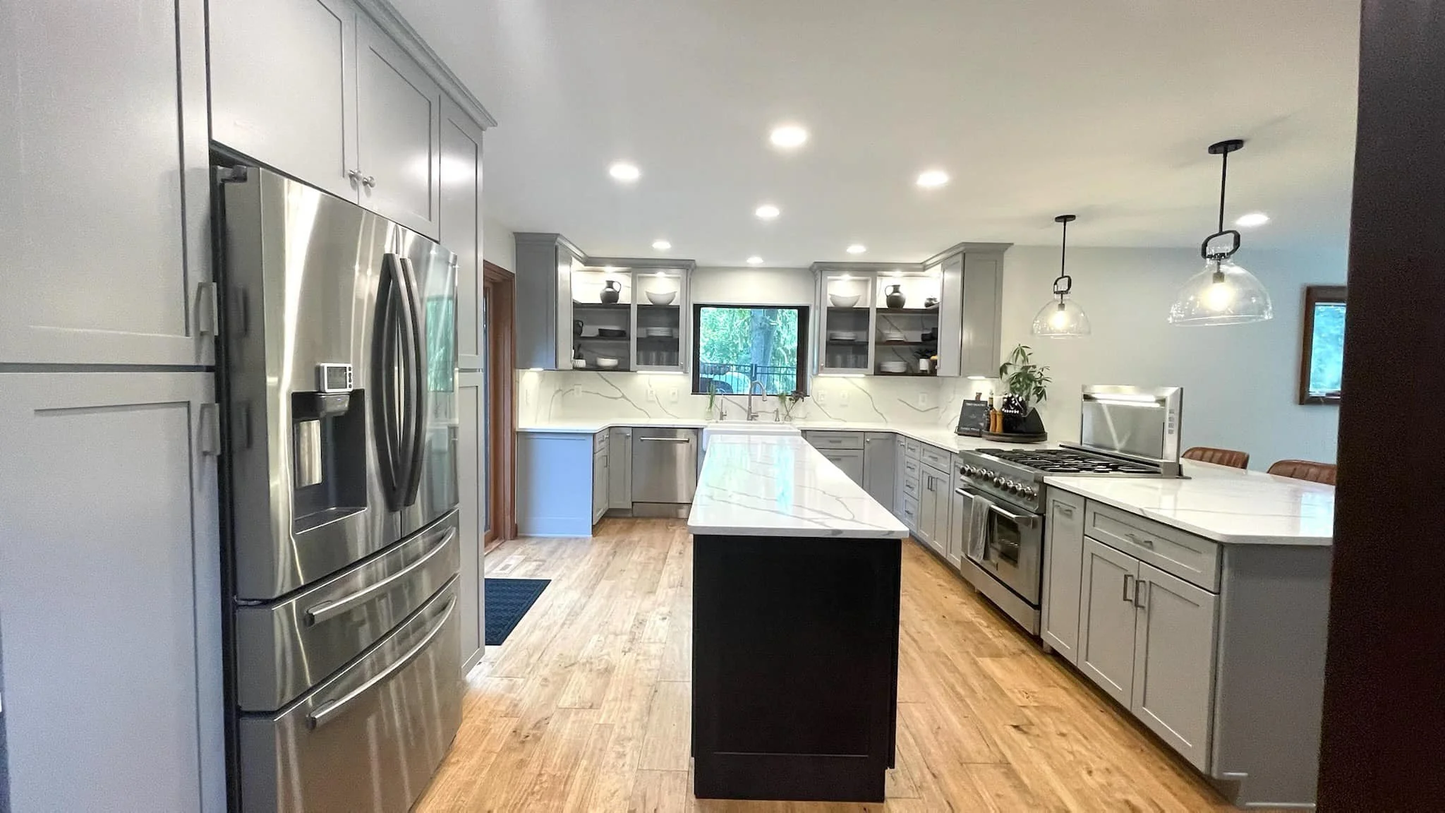 Modern kitchen with gray cabinets, white marble countertops, stainless steel appliances, a central island, pendant lights, and a window overlooking greenery.