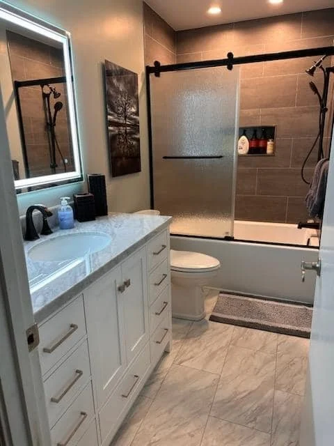 Bathroom featuring a white vanity with a marble countertop, a mirror with lighting, a toilet, a bathtub with a frosted glass sliding door, gray wall tiles in the shower area, a small gray bath mat, and a decorative painting on the wall.