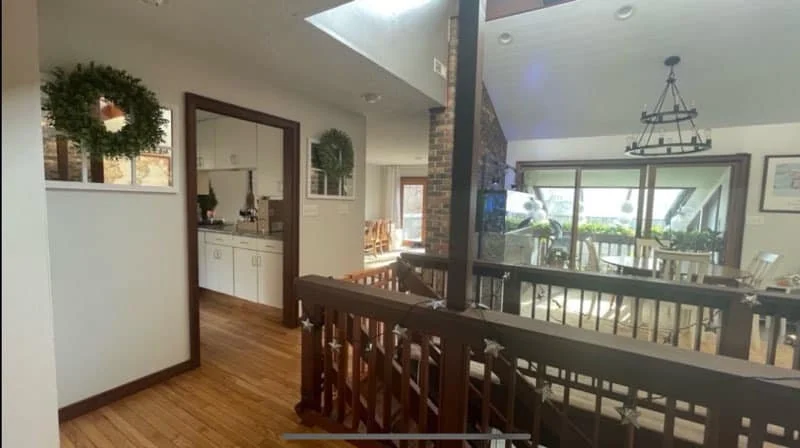 Interior of a house showing a living or dining area with a wooden railing, a dining table, chairs, large sliding glass doors leading to a porch, and decorative wreaths on the wall.