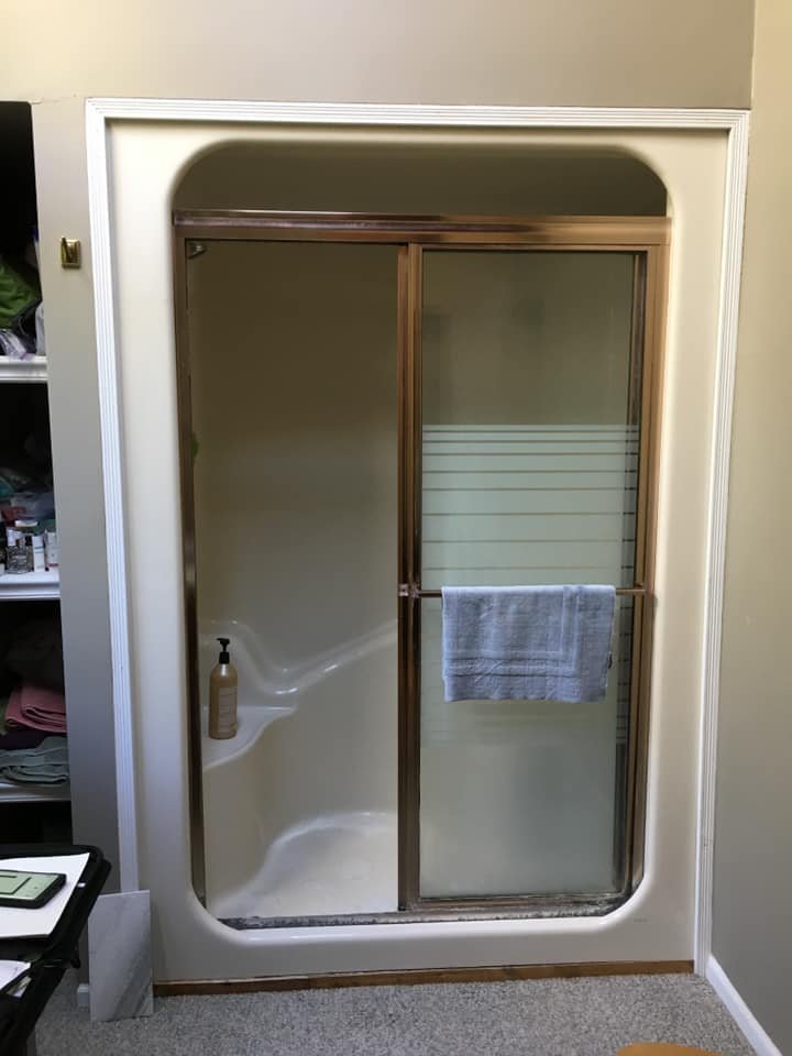 A bathroom shower with a sliding glass door, brass frame, and a built-in soap holder with a bottle of soap or shampoo inside. There is a towel hanging on the door, and a shelf with toiletries in the background.