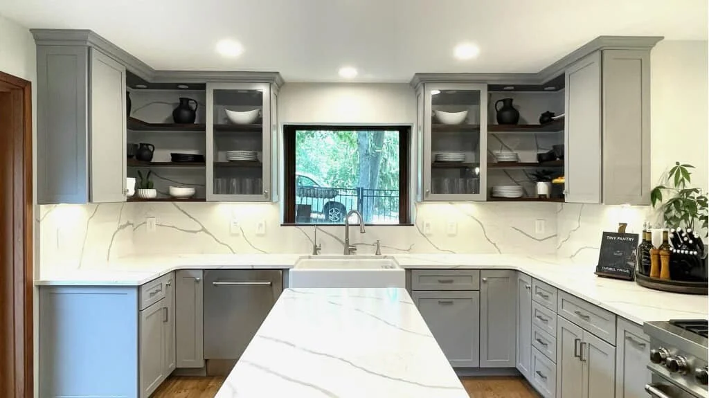 Modern gray kitchen with white marble countertops, a window above the sink, and open cabinets displaying dishes and black pots.