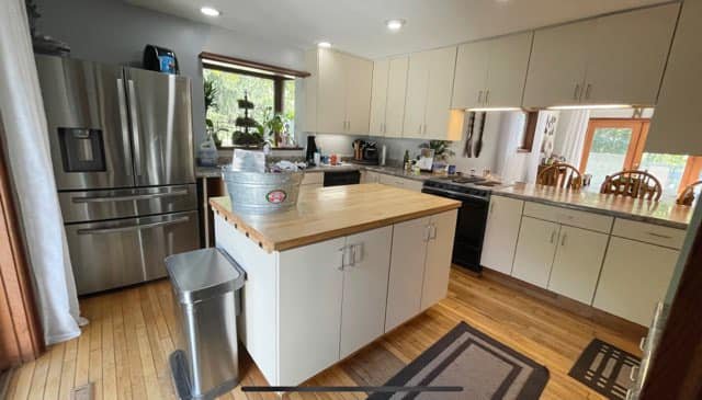 Modern kitchen with white cabinets, stainless steel refrigerator, and a wooden island countertop. There is a window with a plant, and a dining area with chairs and a large window in the background.