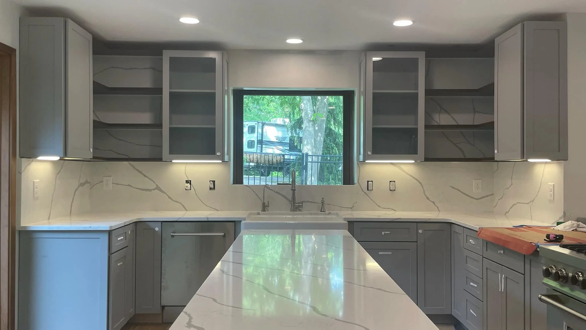 Modern kitchen with gray cabinets, marble backsplash, and a large central island, featuring a window above the sink showing trees and parked RV outside.
