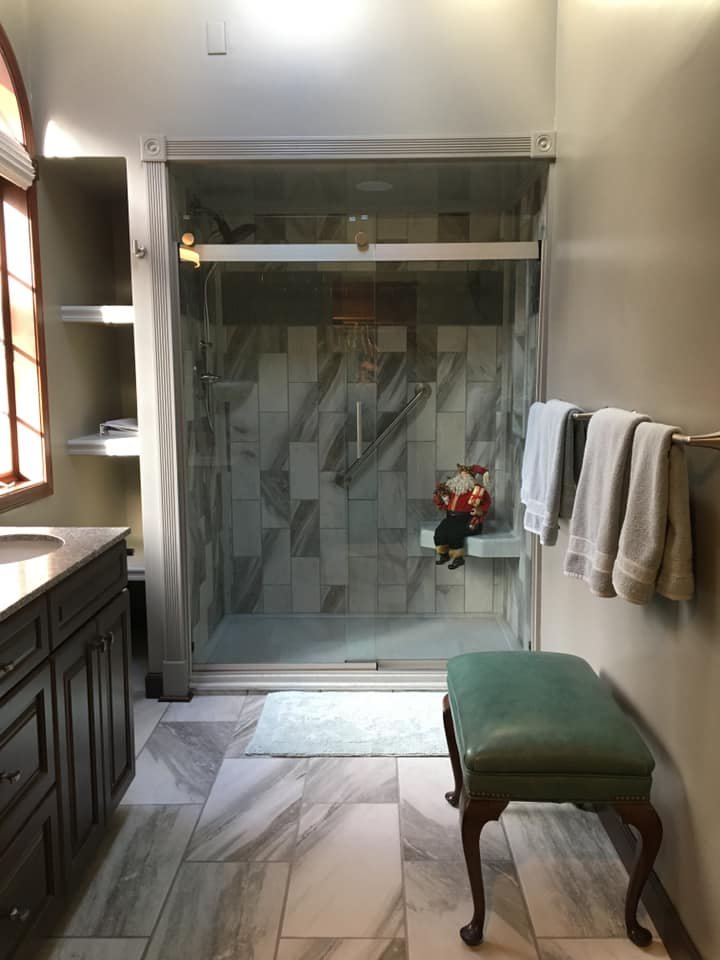 Bathroom with a glass shower stall, a green cushioned stool, and towels on a rack. There is a toy Santa Claus figure seated on the shower bench.