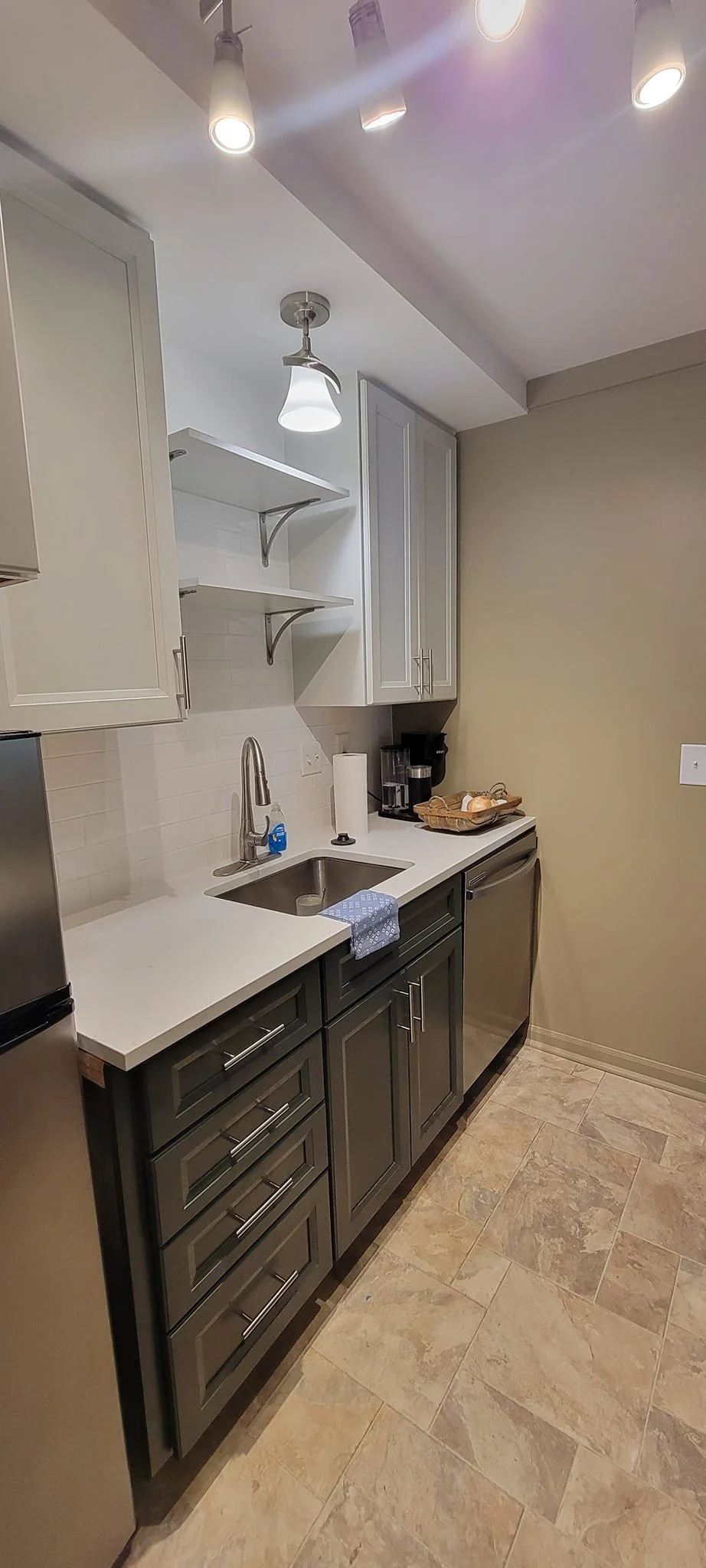 A small kitchen with gray cabinets, a white countertop, a stainless steel sink, a paper towel roll, coffee maker, and open shelving, under ceiling lights. 