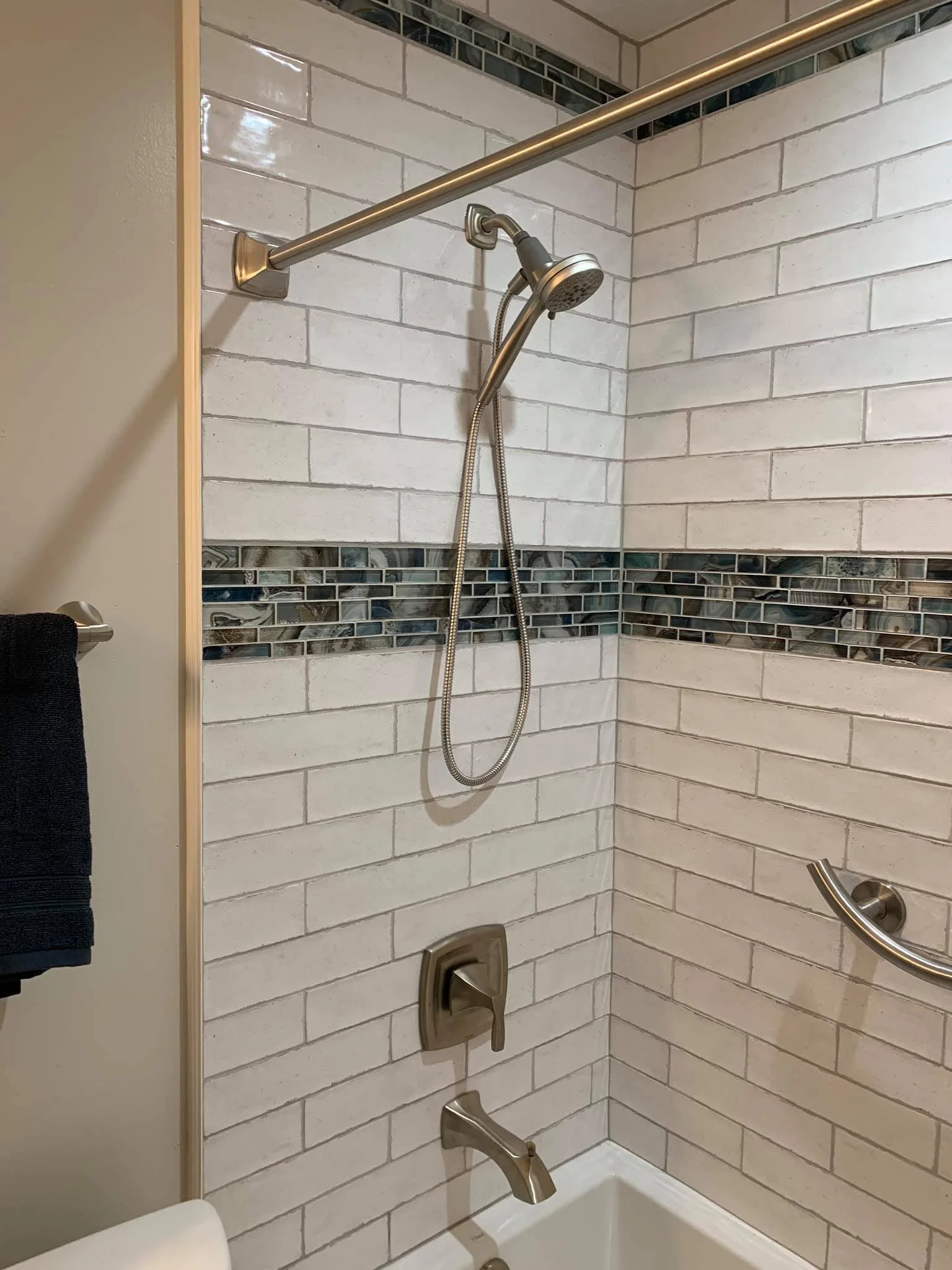 A bathroom shower with white tile walls, a decorative tile border, a brushed nickel showerhead, a hand-held shower spray, a horizontal grab bar, and a wall-mounted faucet.