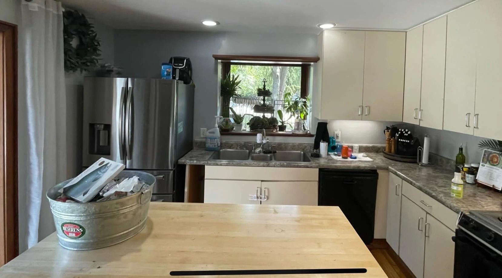 Kitchen with stainless steel refrigerator, beige cabinets, granite countertops, window with plants, multiple small kitchen appliances, and a wooden island in the foreground.