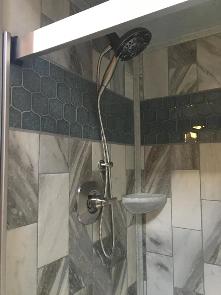 A close-up of a modern shower corner with a black showerhead, a hand-held shower hose, a control knob, a soap dish, and wall tiles in shades of gray and white.