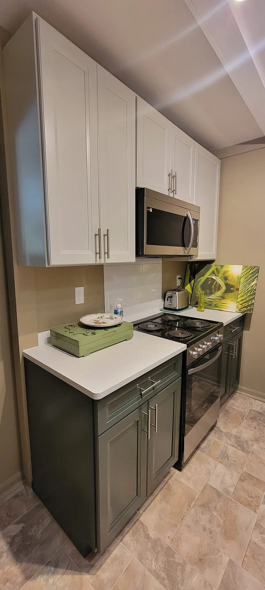 Small kitchen with white upper cabinets, dark lower cabinets, a white countertop, a stove, microwave, toaster, and decorative artwork with green and natural tones.