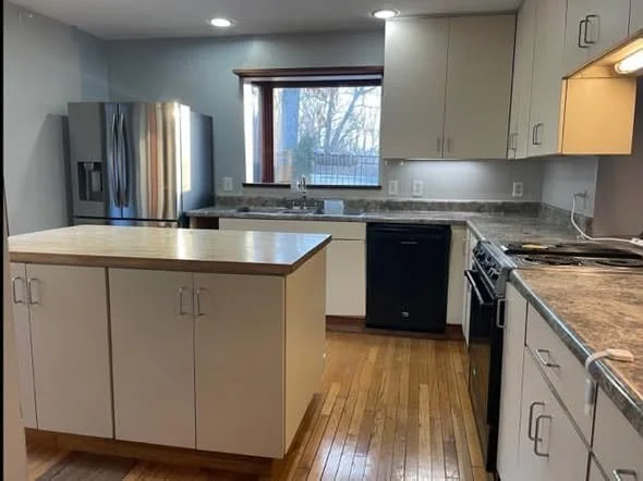 Kitchen with white cabinets, granite countertops, stainless steel refrigerator, black dishwasher, oven, and kitchen island, with a window showing trees outside.