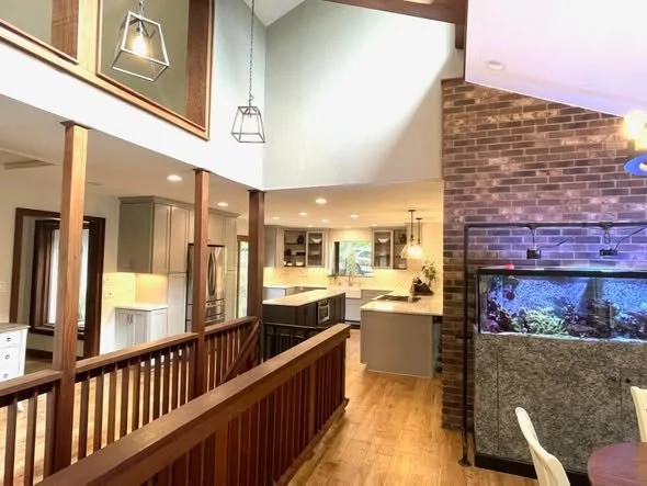 Interior of a modern house showing a kitchen with white cabinets, a large window, and a central island, adjacent to a living or dining area with a brick wall and a large fish tank.