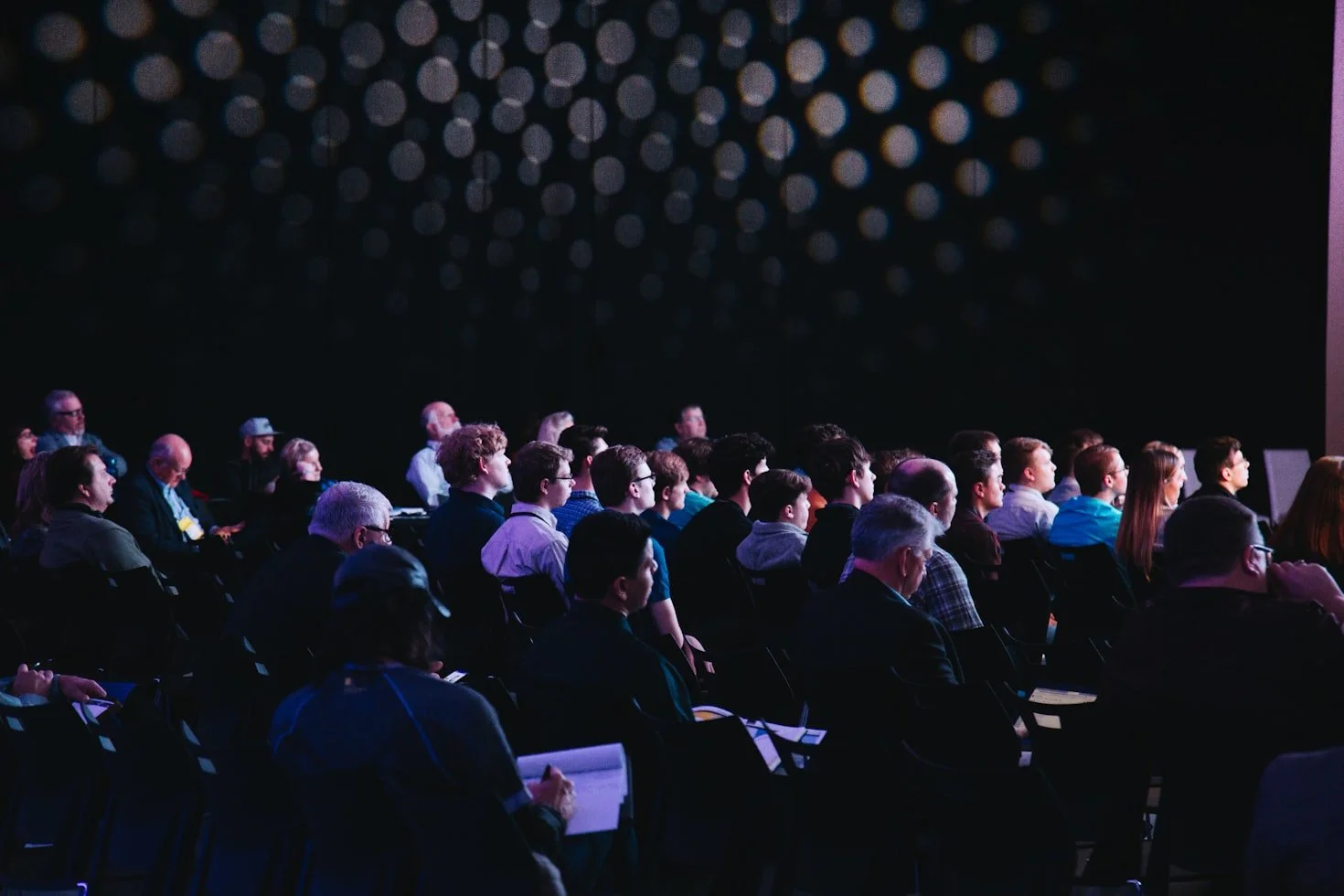 Audience seated in a darkened conference room, watching a presentation or speaker.