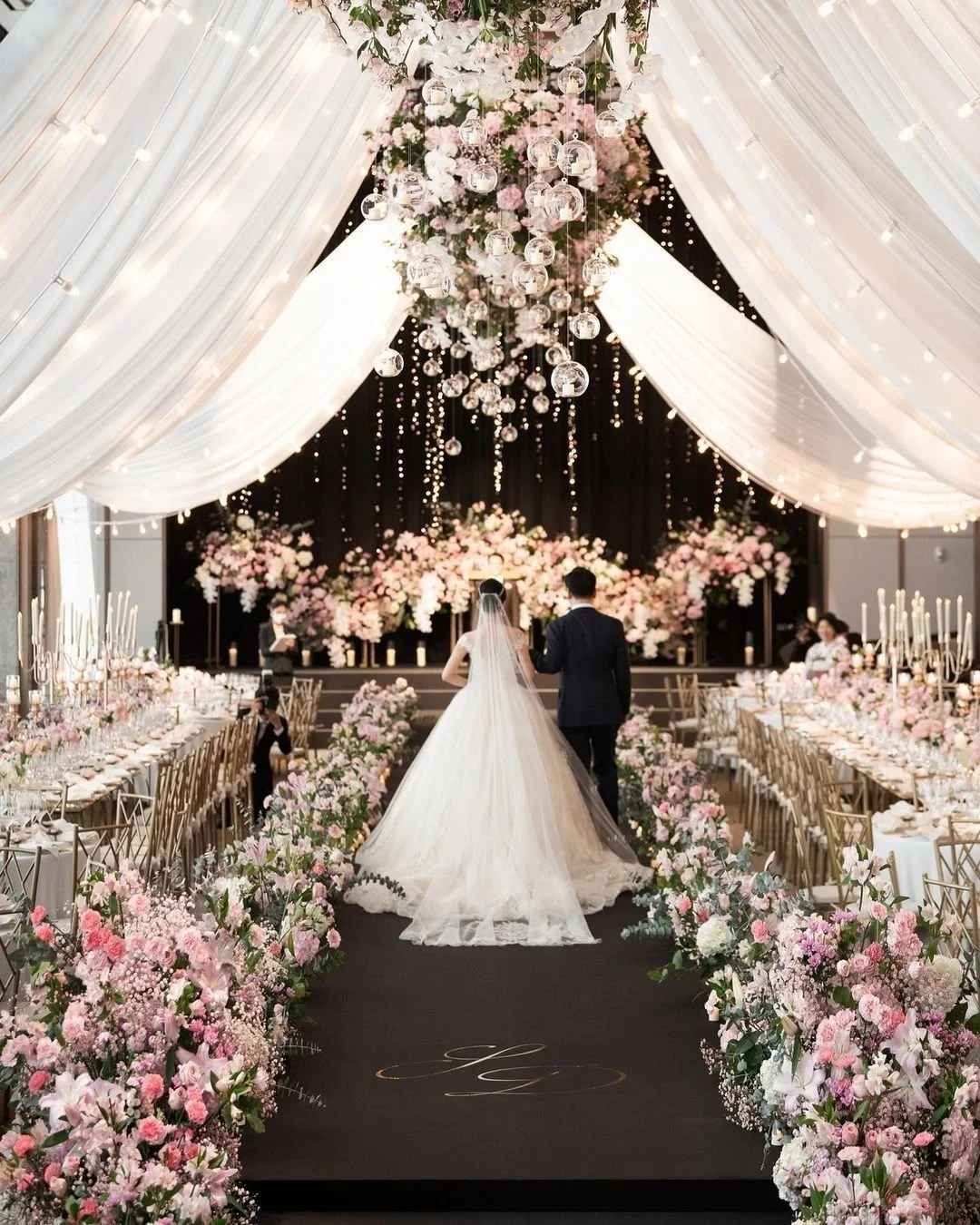 A wedding ceremony with a bride and groom walking down the aisle inside a decorated venue with pink and white flowers, draped white fabric, and hanging glass globes.