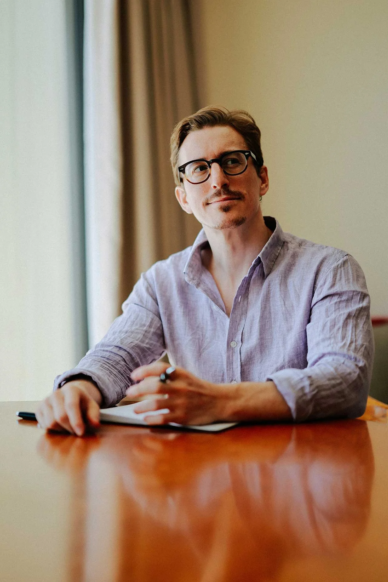 A man sits at a desk, holding a pen and notebook, and looks thoughtfully away from the camera