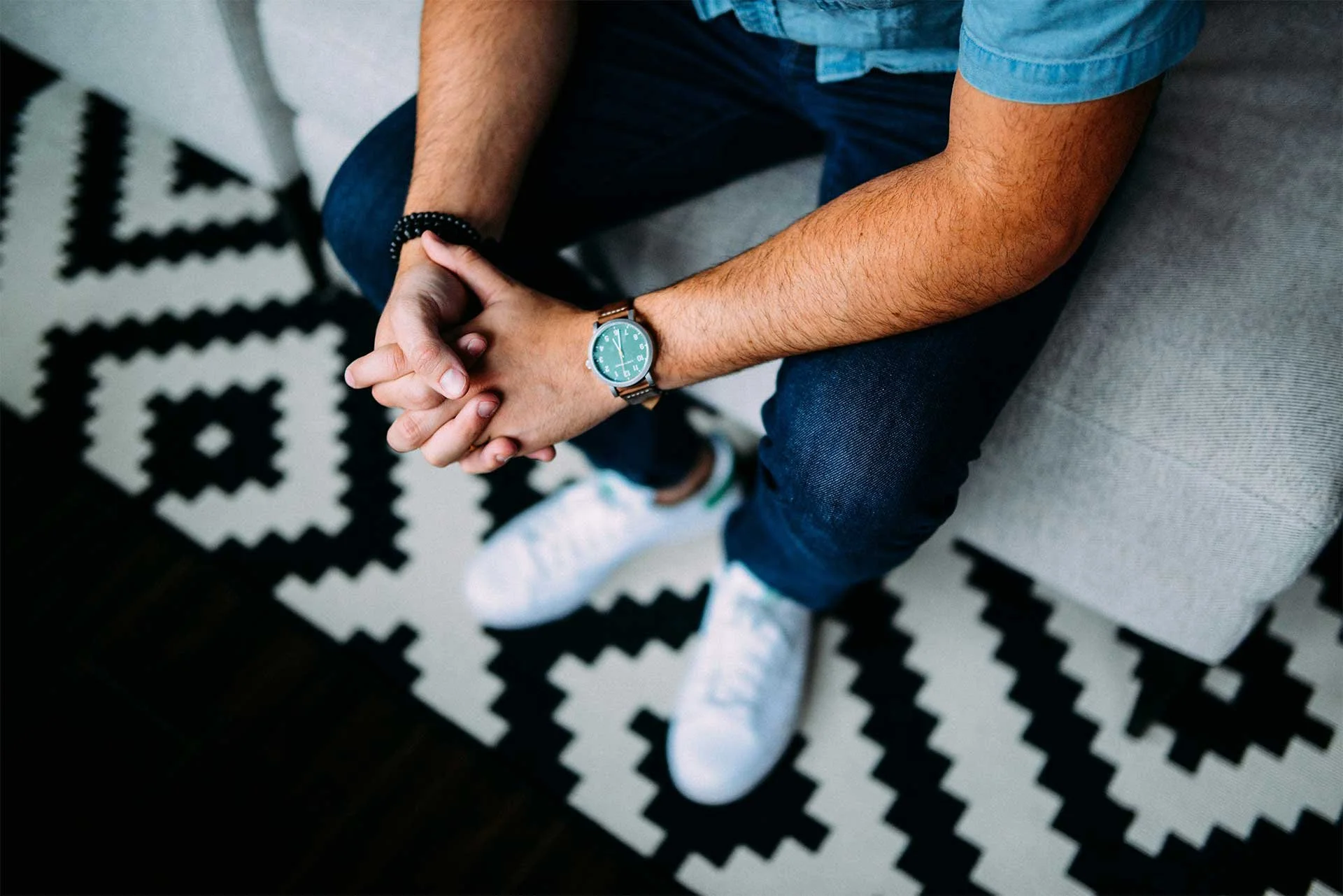 A man seated at a couch leaning forward with hands clasped, wearing a watch, jeans, and white sneakers.