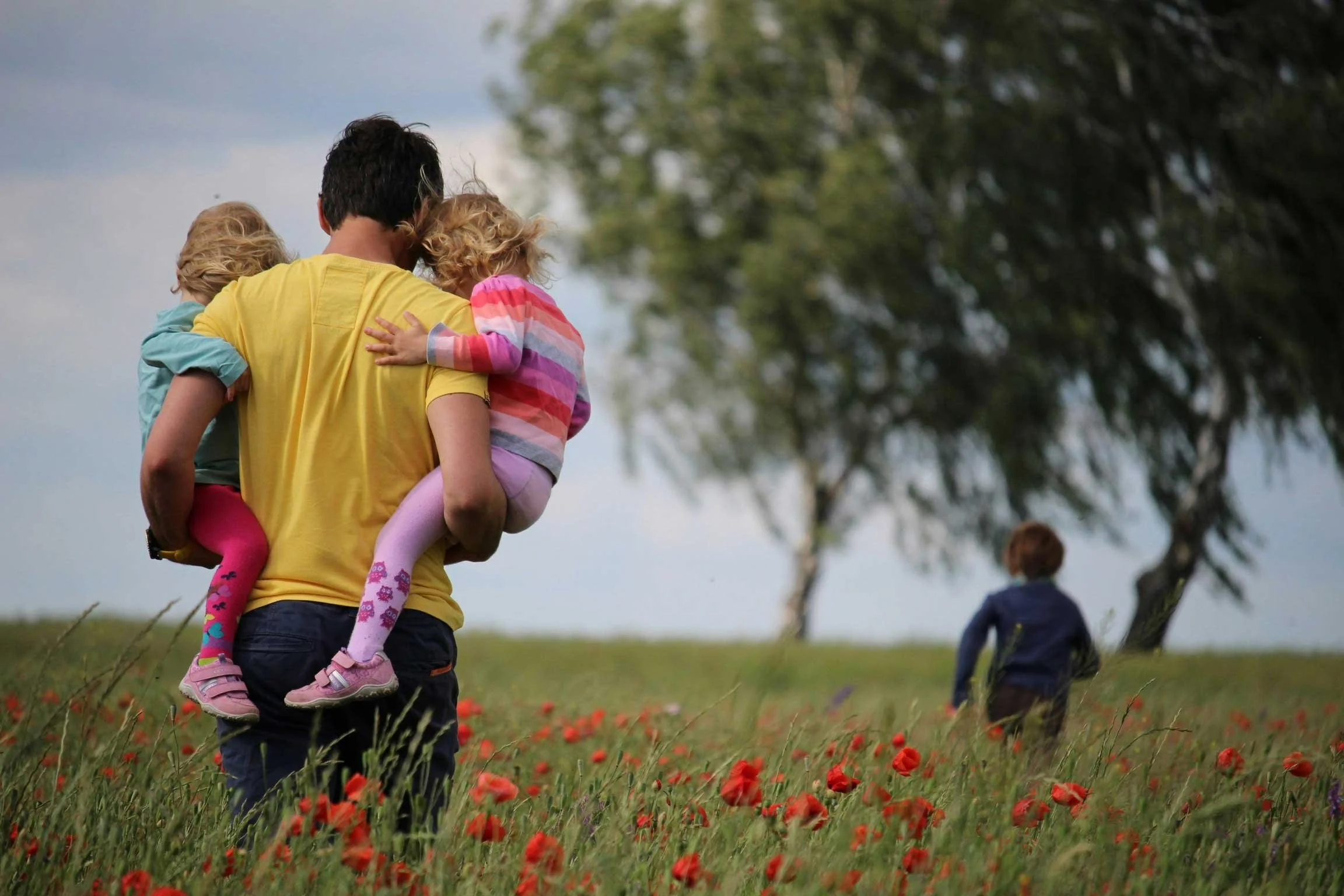 Single dad holding one daughter in each arm, facing away from us and standing outside.