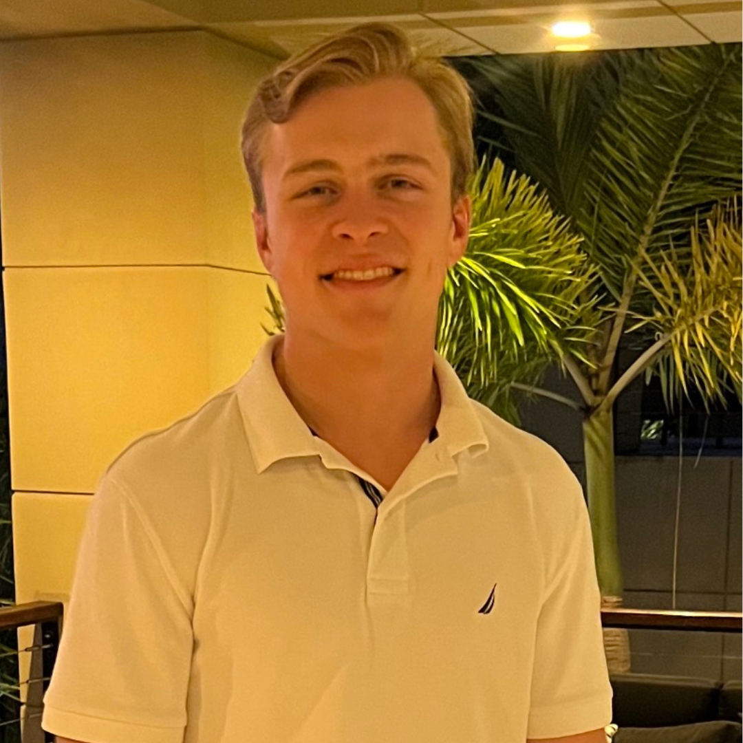 Person wearing a white polo shirt standing indoors with palm trees in background.