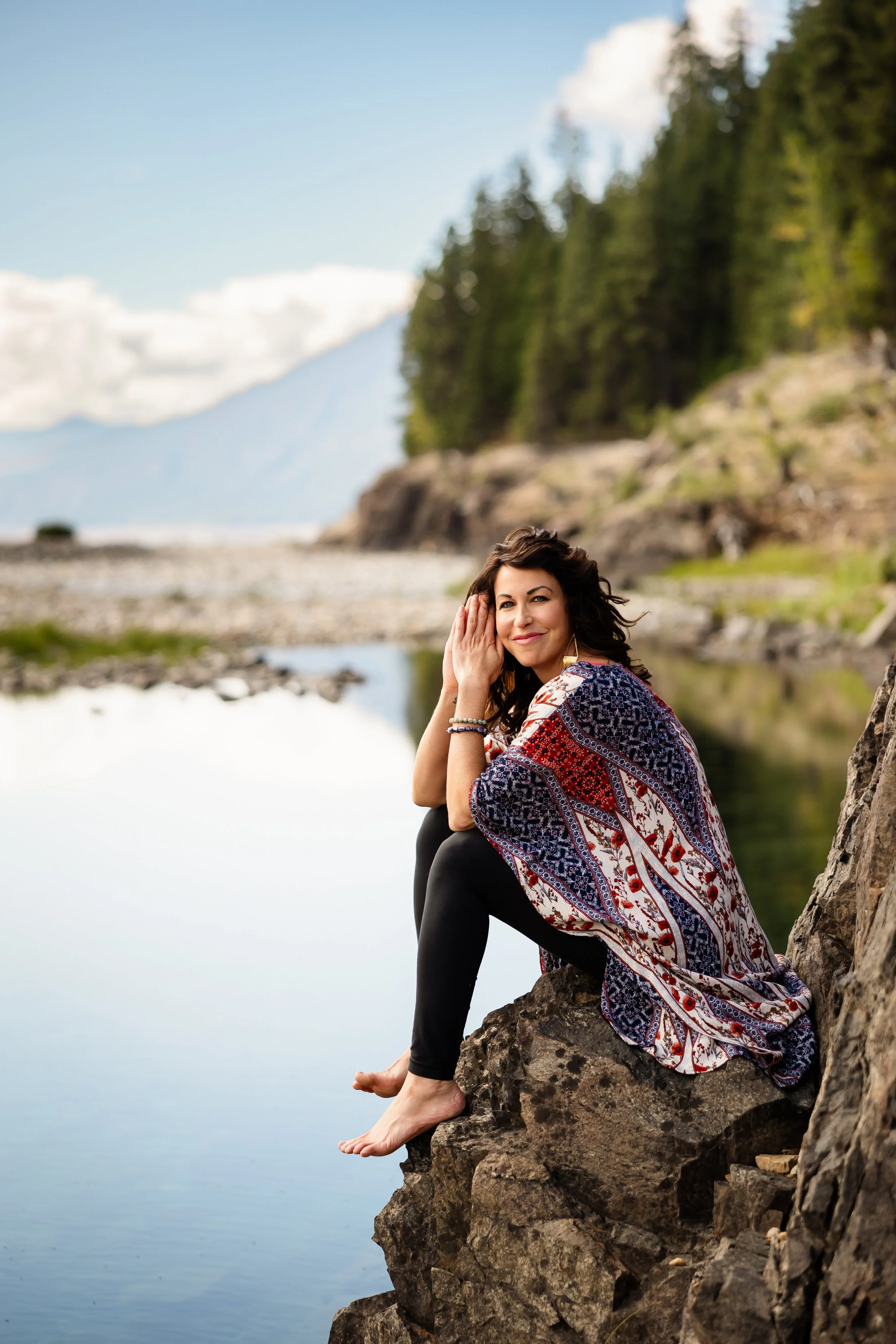 Woman seated on rocks by a lake, wearing a colorful patterned shawl, with trees and mountains in the background.