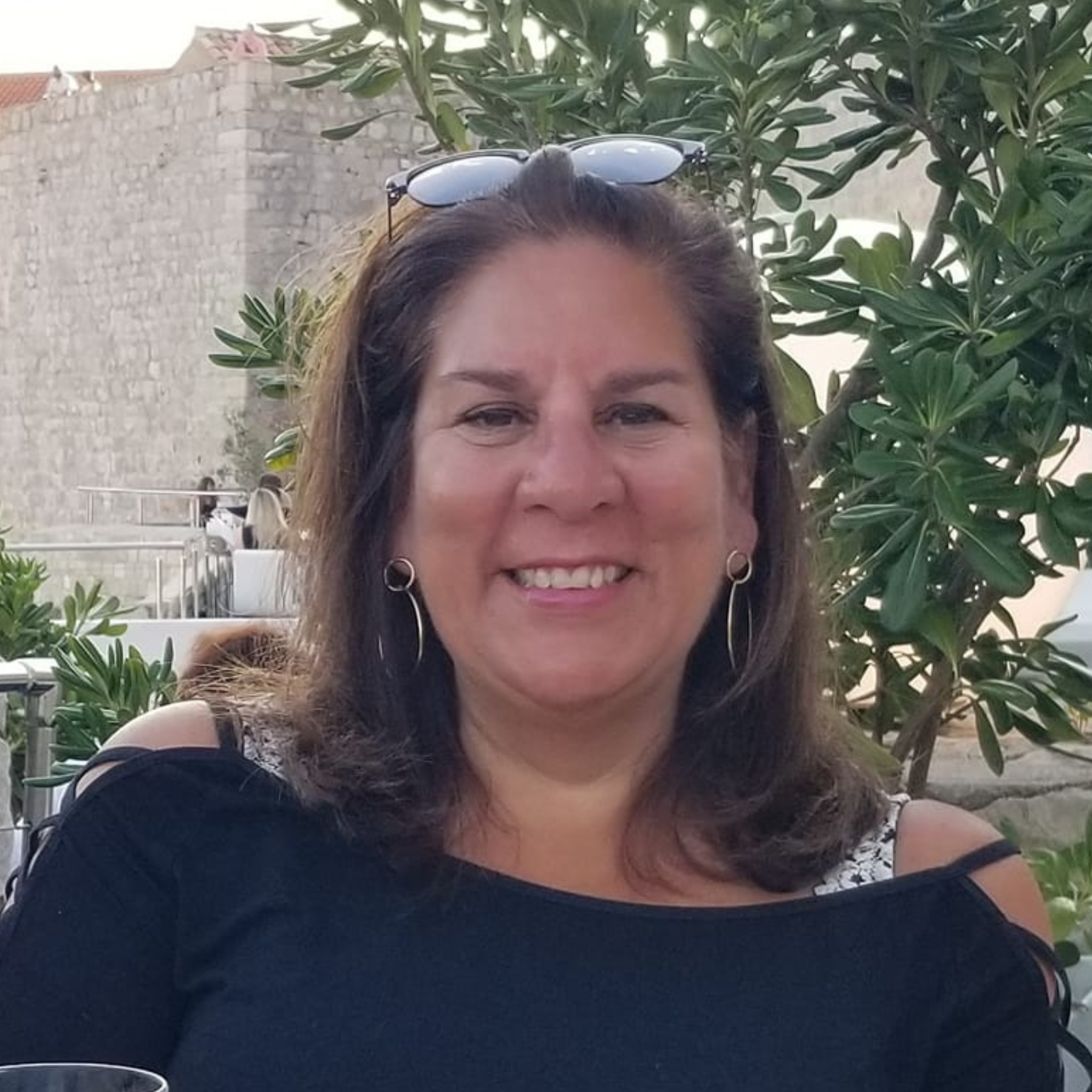 Smiling woman wearing sunglasses on her head, posing outdoors with a background of greenery and a stone wall.