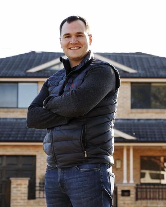 A man standing outside in front of a house with crossed arms, smiling, wearing a black puffer vest and jeans.