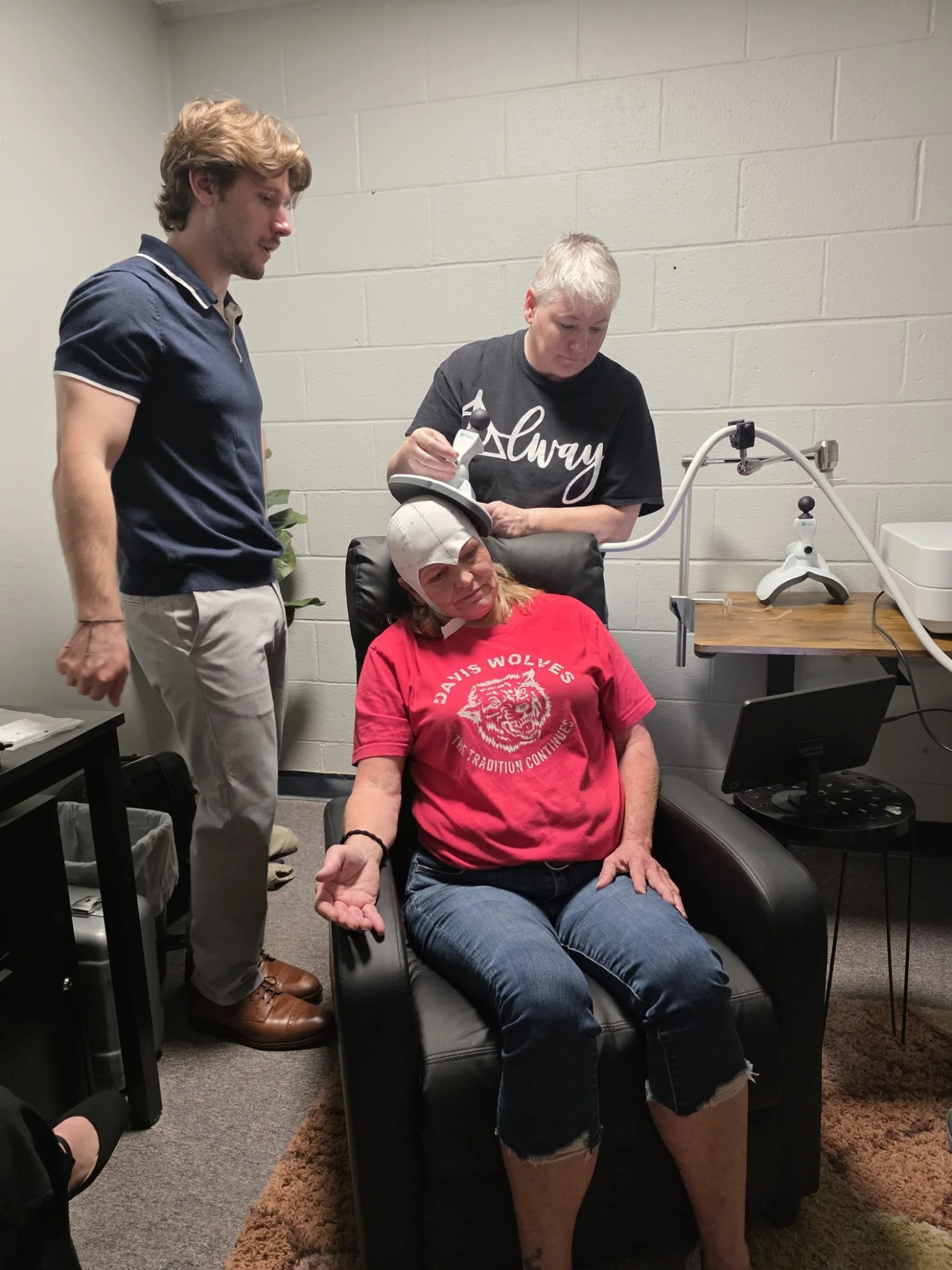 Two people assist a seated woman wearing a head cap during a medical examination in a clinic room.